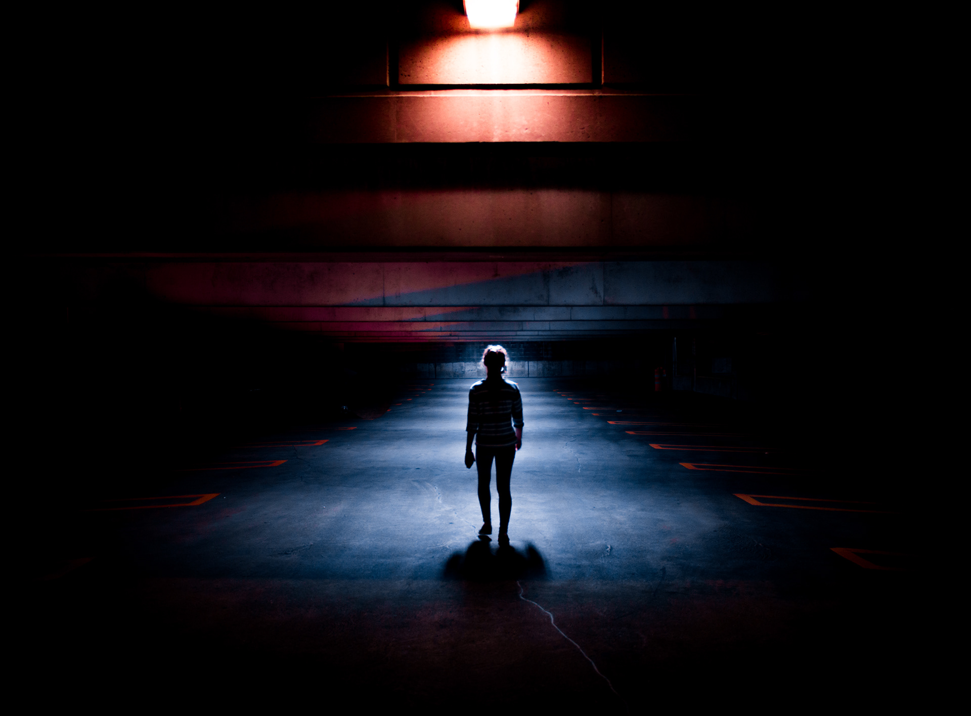 Person standing alone in a dark, empty space illuminated by a bright light above, with a shadow on the ground.