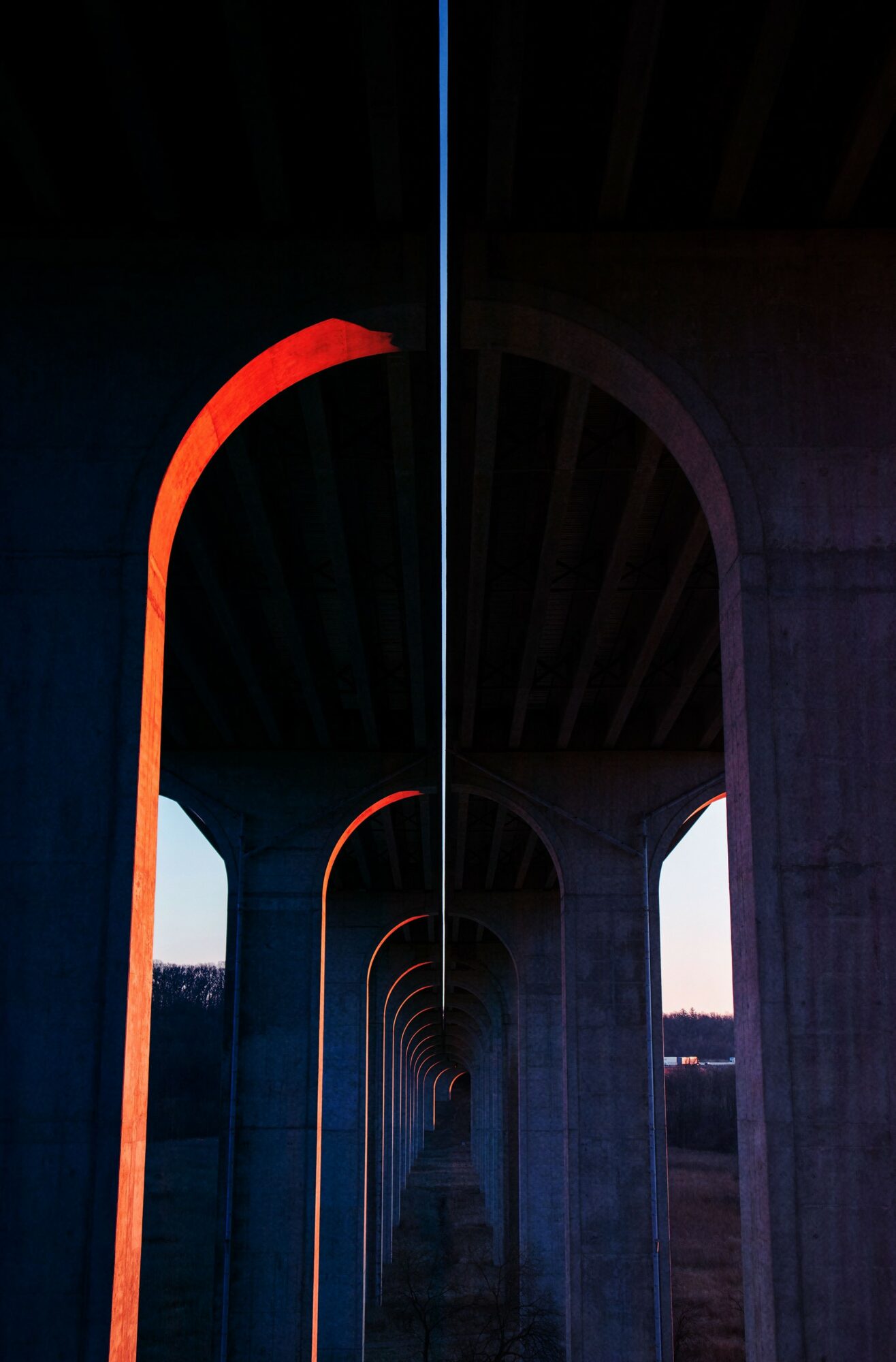 Under a bridge with illuminated arches, view down a tunnel-like structure with colorful lighting, leading to a bright opening.