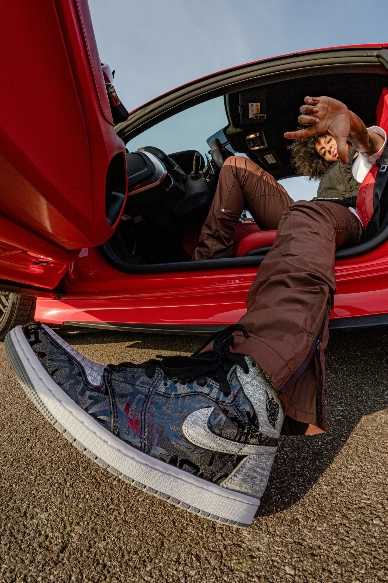 Person sitting in a red car with one leg outside, wearing sneakers and brown pants, holding a phone.