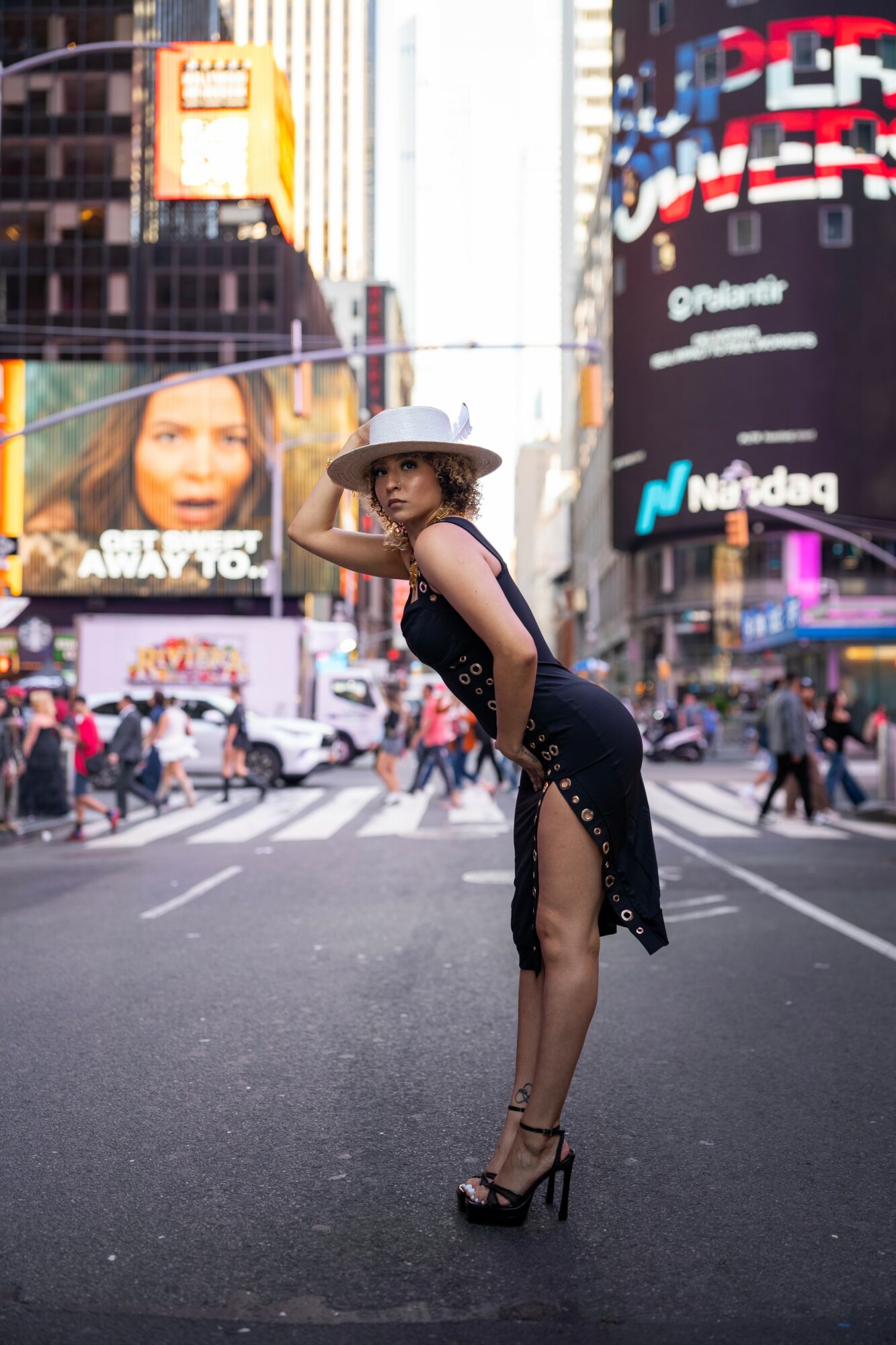 Woman in black dress and hat standing in city street with digital billboards and pedestrians in background.