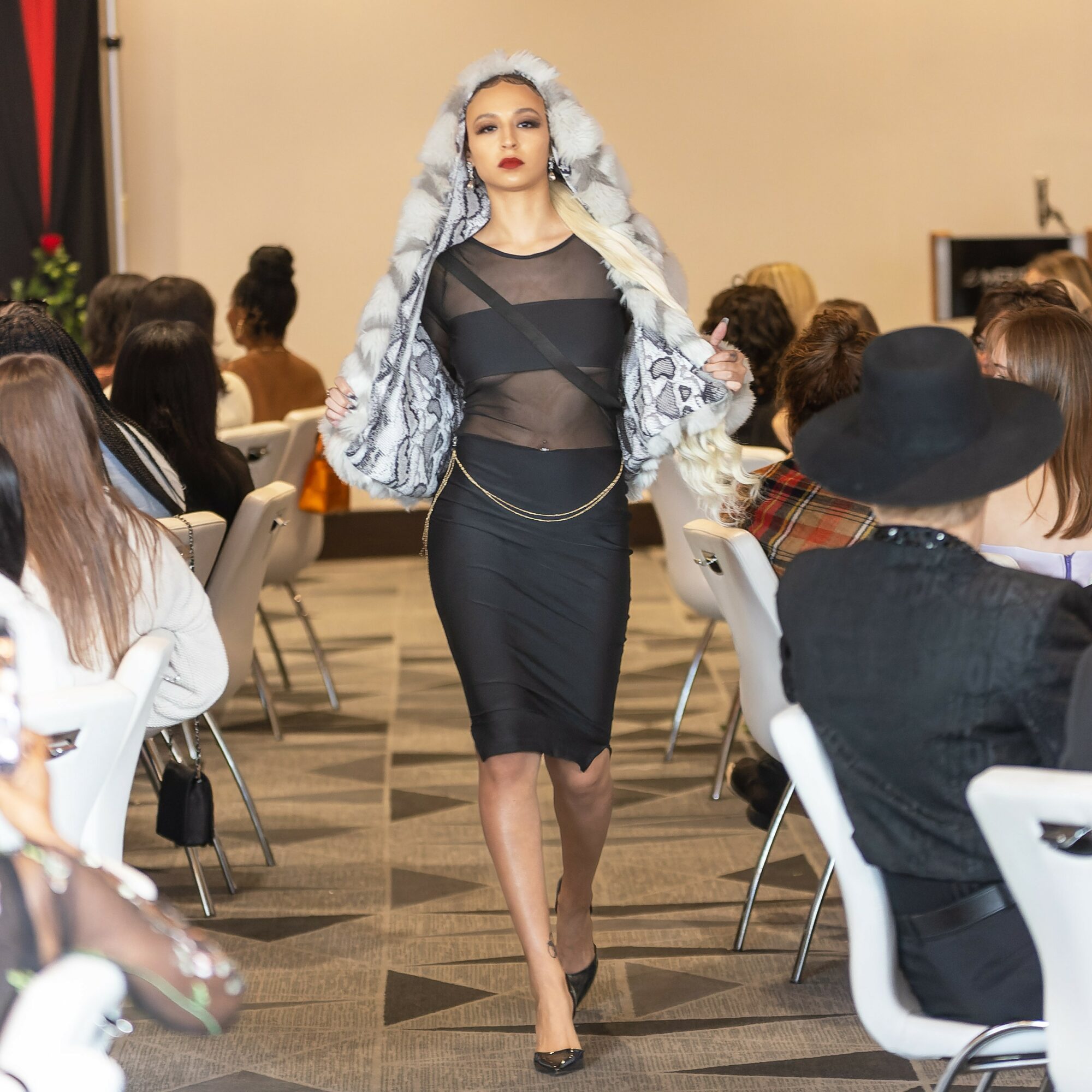 A woman walks down a runway in a black dress with a sheer top and a large patterned headscarf, audience seated on both sides.
