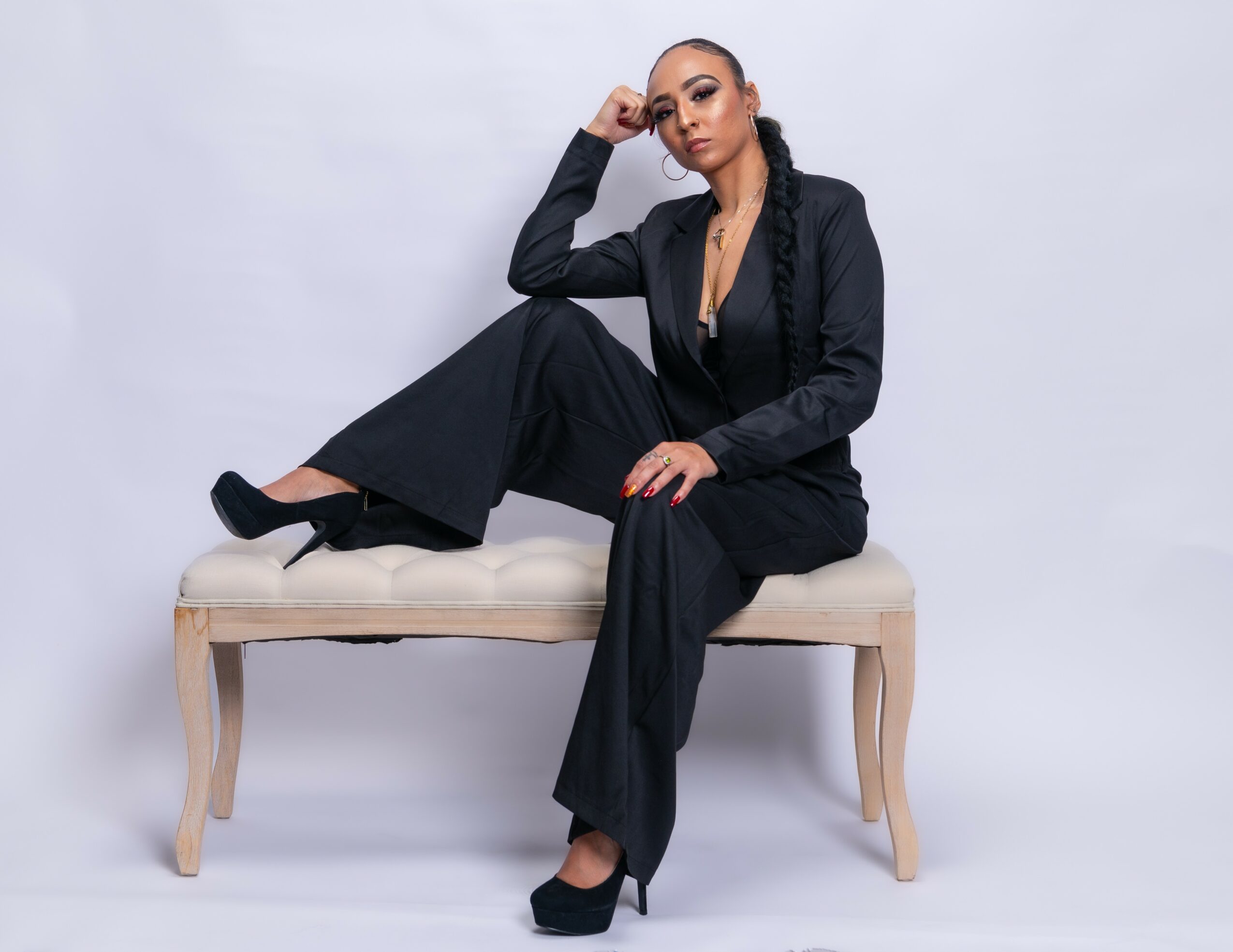 Woman in black suit sitting on a bench, one leg crossed over the other, posing confidently against a plain background.