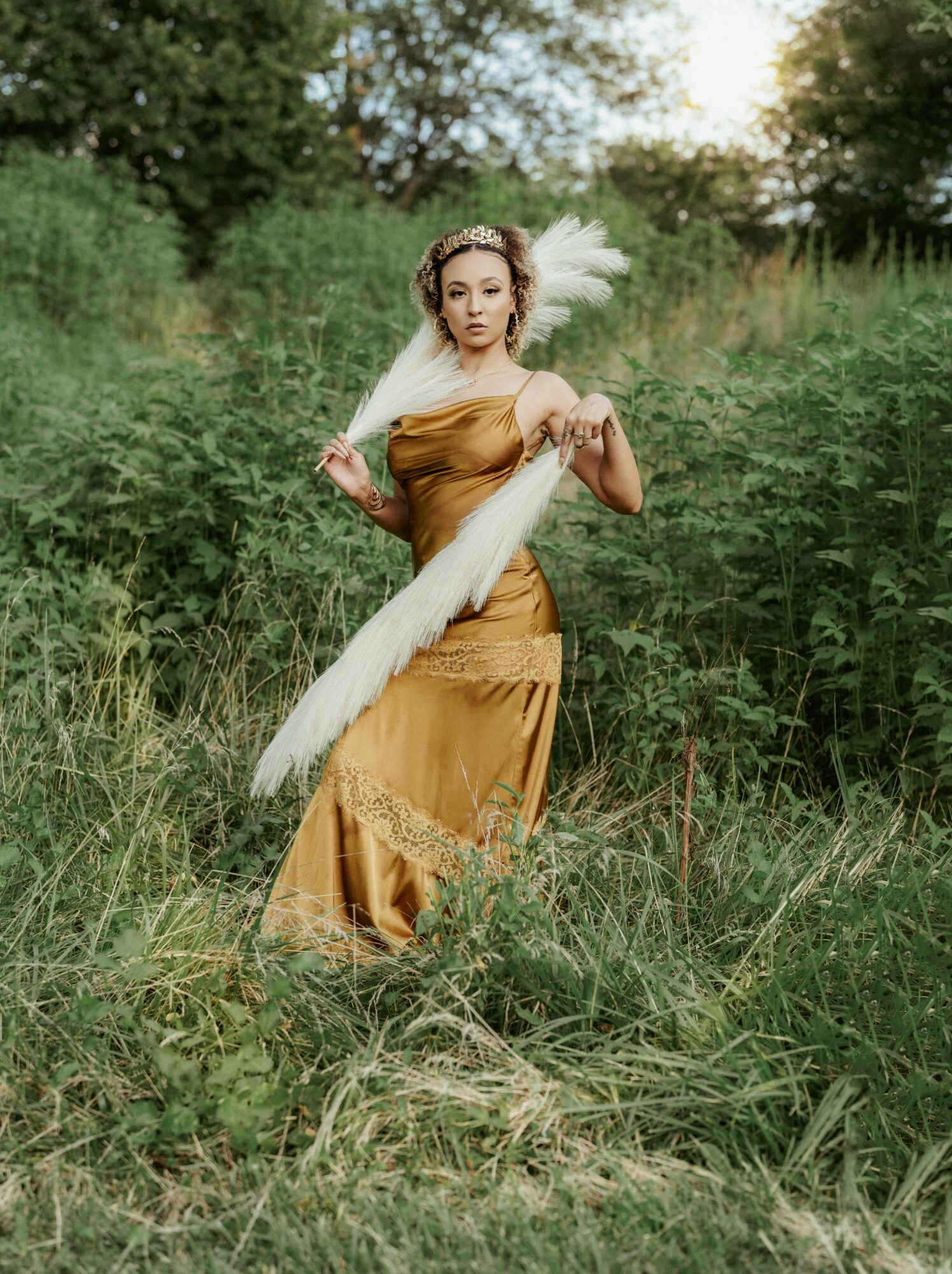 Woman in a gold dress holding a large white feather in a grassy field with trees in the background.