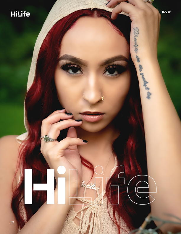 Close-up portrait of a woman with red hair, piercing eyes, and tattoos, outdoors with a blurred green background.
