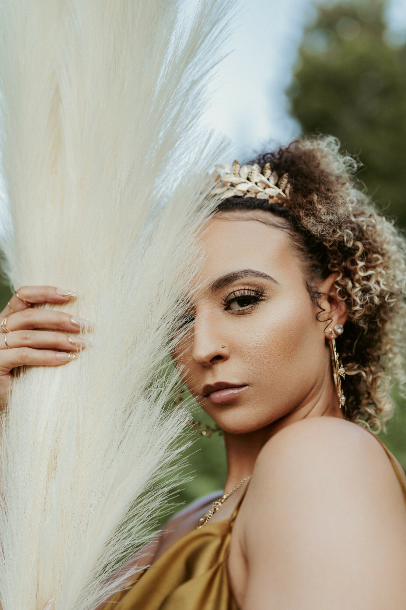 Young woman with curly hair and headband, partially hidden behind tall white grass outdoors.