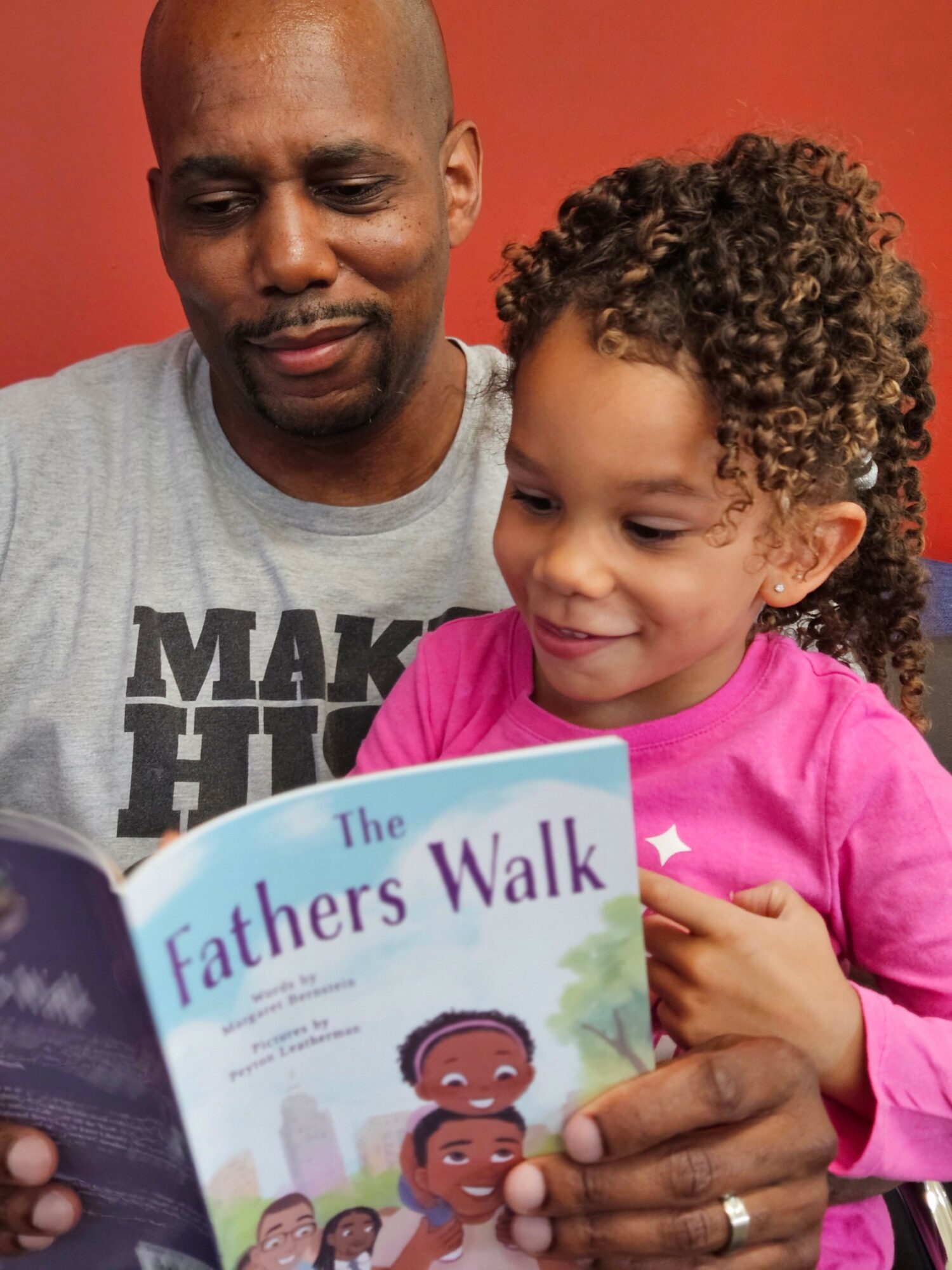 A young girl and an adult man look at a pamphlet together, smiling, against a red background.
