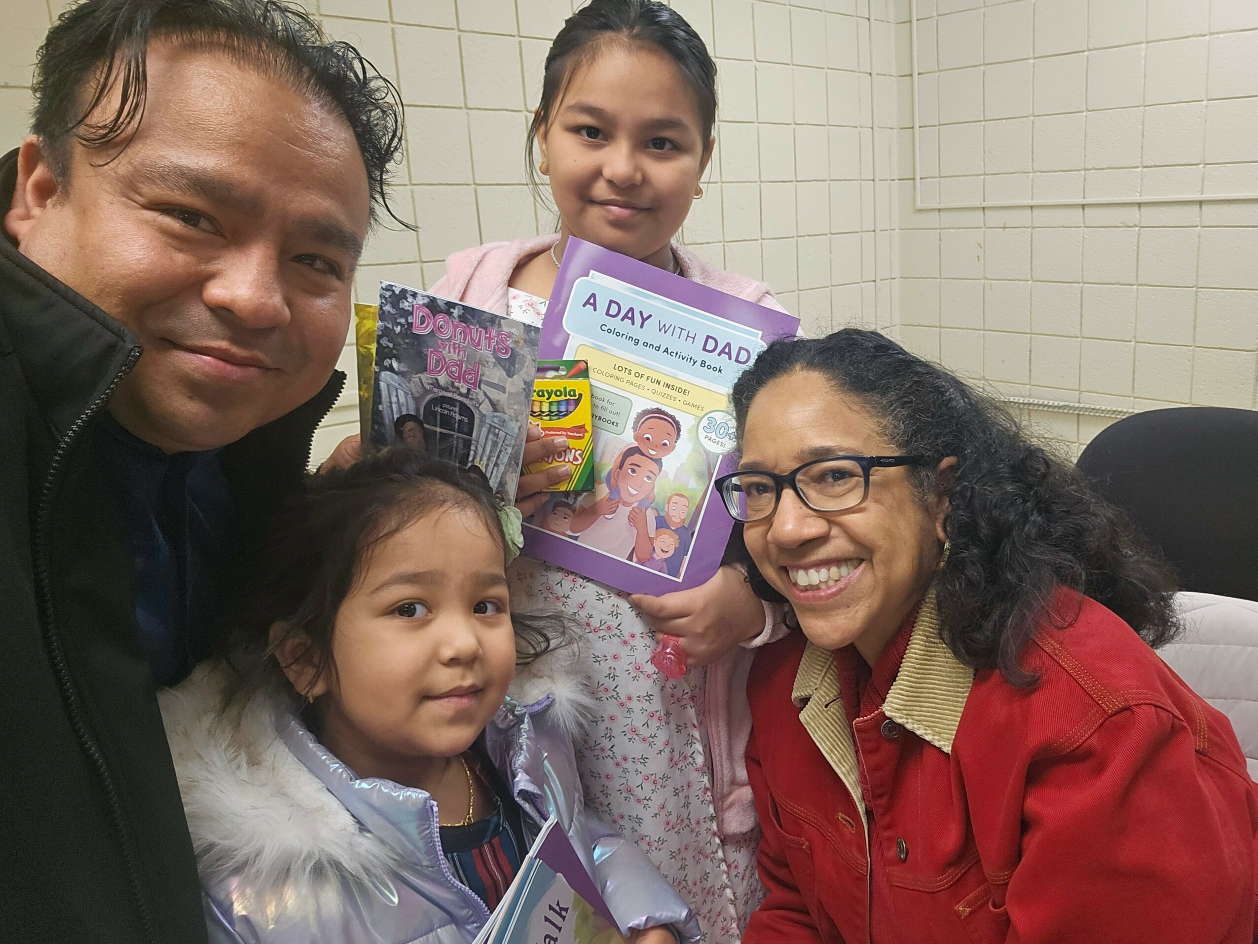 Four people, two adults and two children, smiling indoors with tiled walls, holding a gift box and snacks.