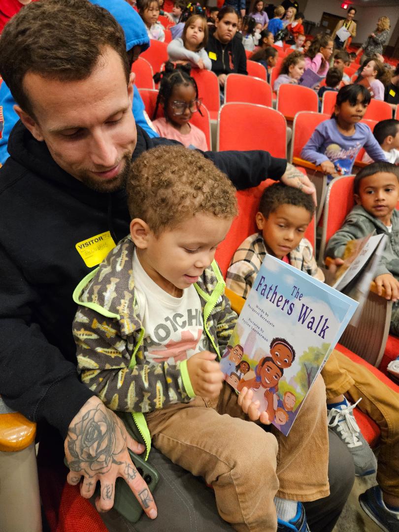 Adult and child reading a book titled 'Father's Walk' in a crowded auditorium with children seated in red chairs.