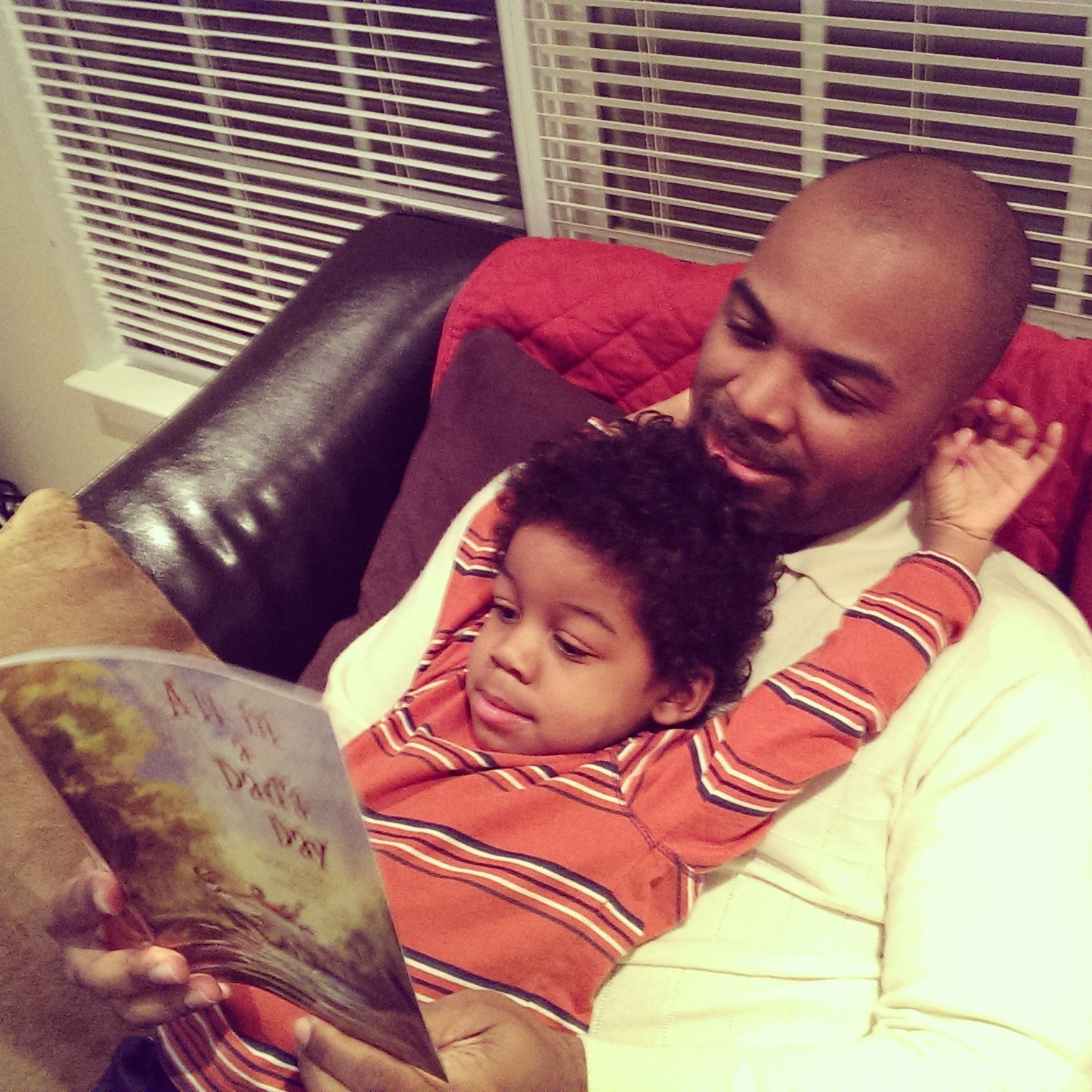 Adult and child sitting on a couch, reading a book together, with a window and blinds in the background.