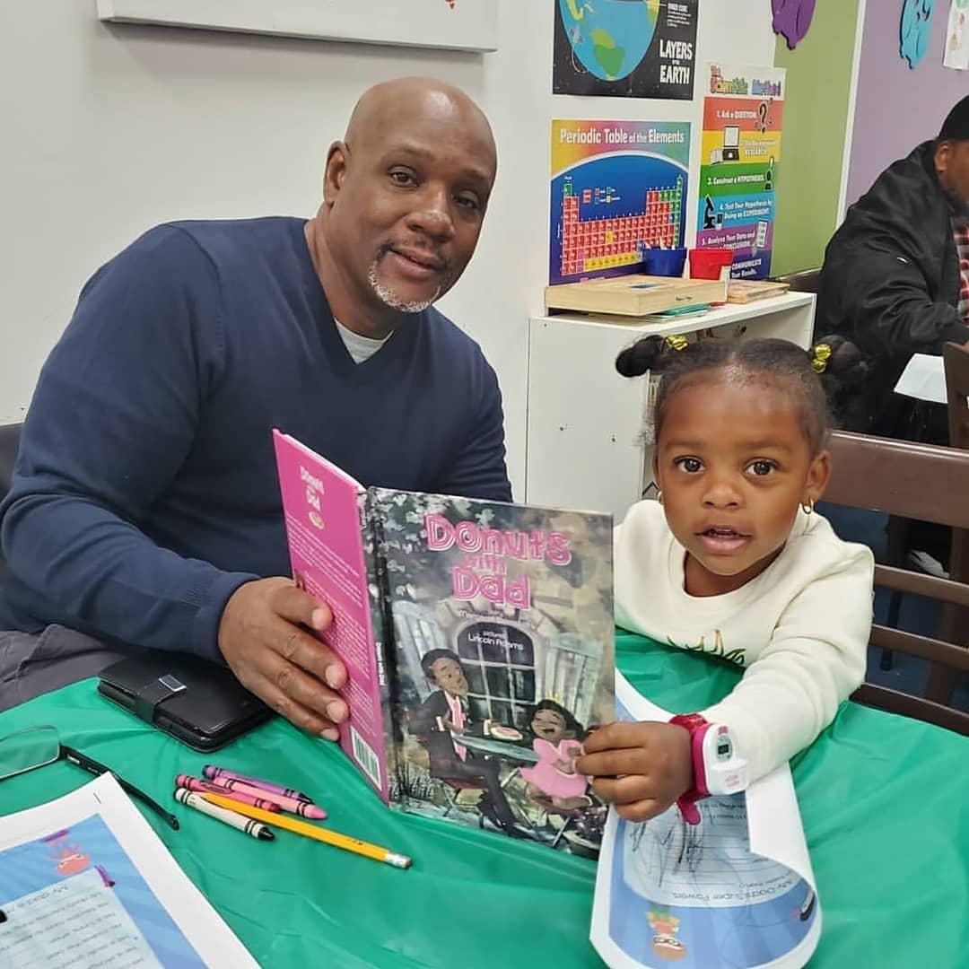 A man and a young girl sit at a table holding a colorful book, with school posters in the background.