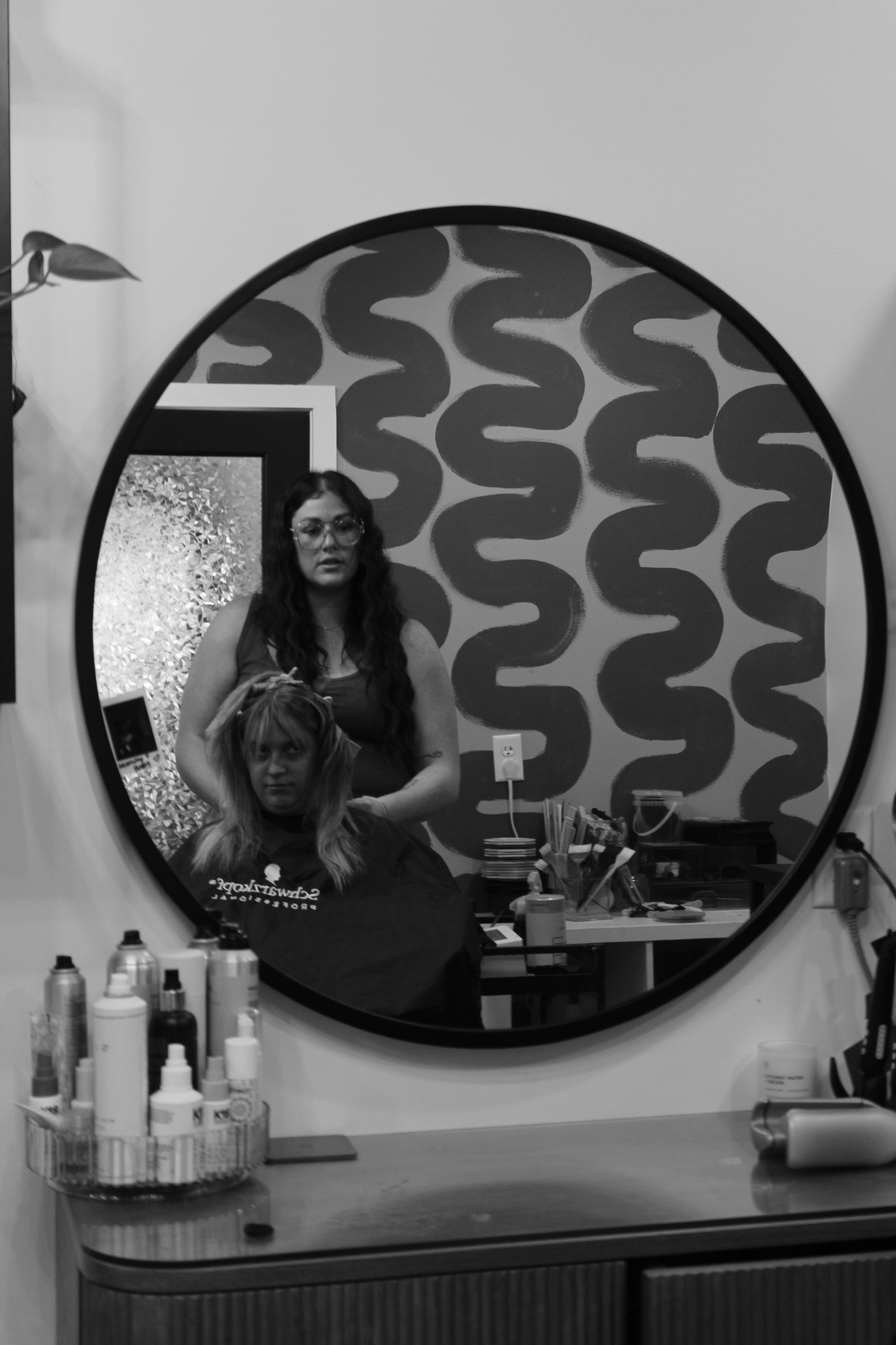 Person taking a mirror selfie in a room with patterned wallpaper, holding a phone, with various bottles on a table.