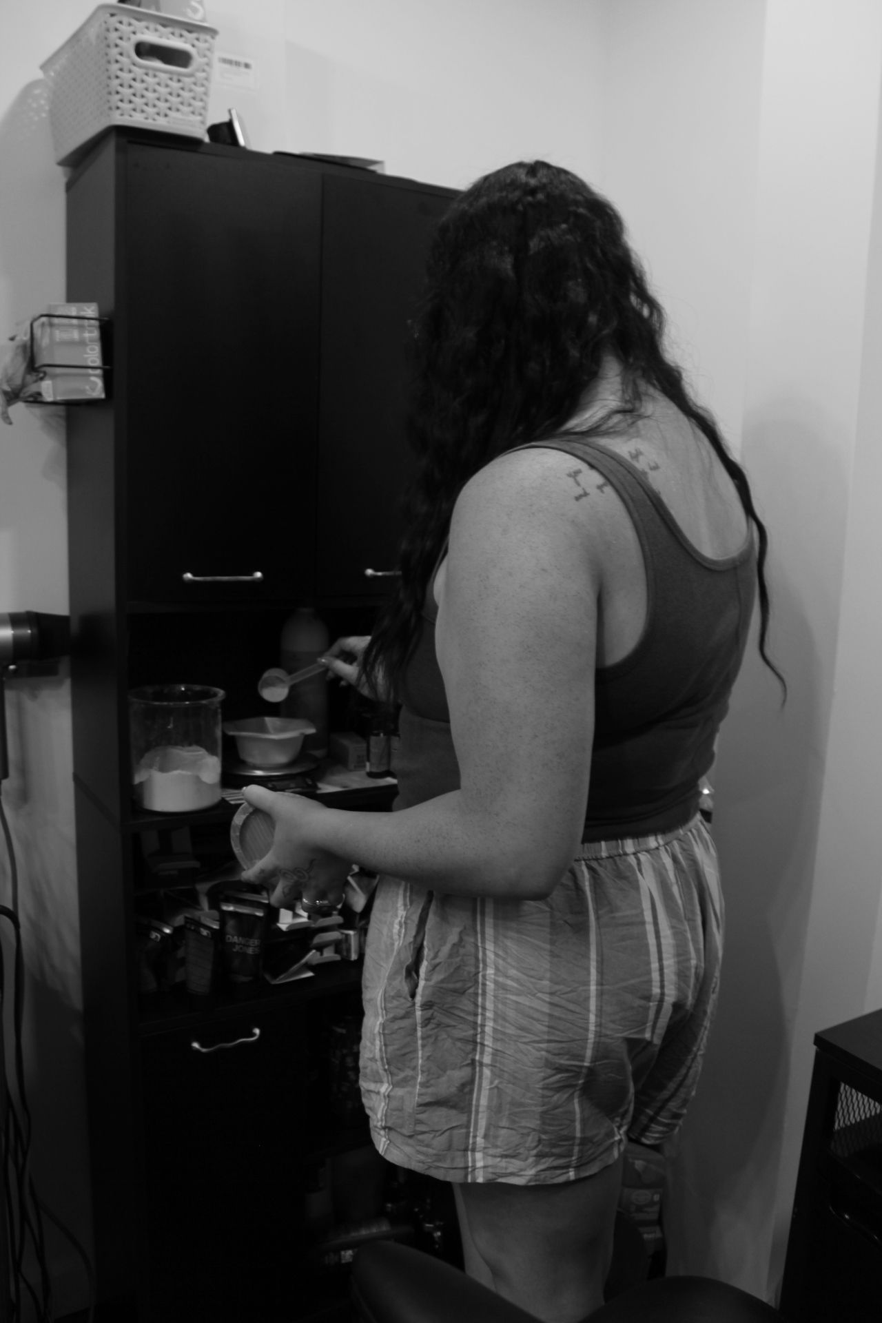 Person with long hair reaching into a kitchen cabinet, wearing a striped shirt and shorts, viewed from the side.