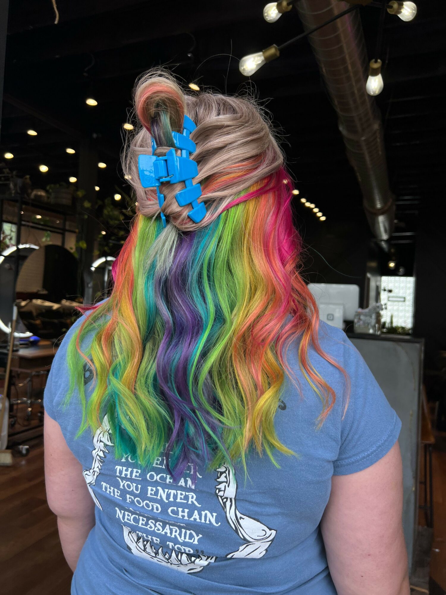 Person with long, rainbow-colored hair wearing a blue T-shirt, indoors with string lights and dark background.