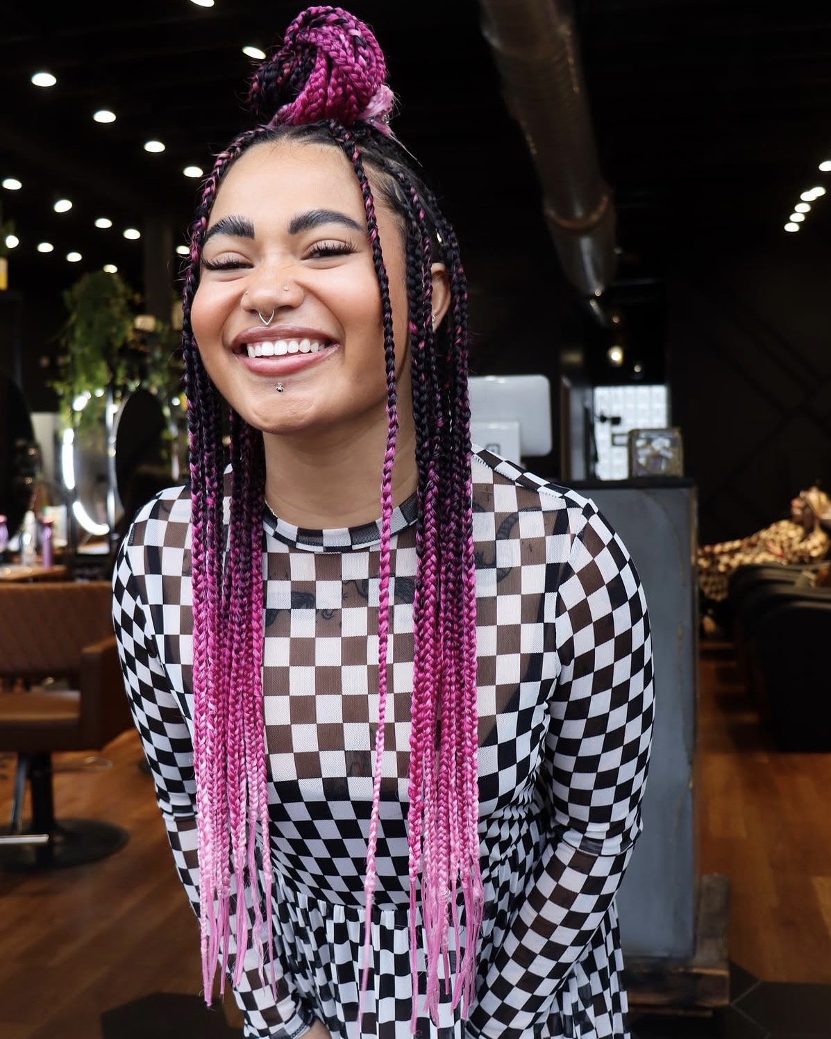 Smiling woman with pink braided hair and checkered top in a modern indoor setting.