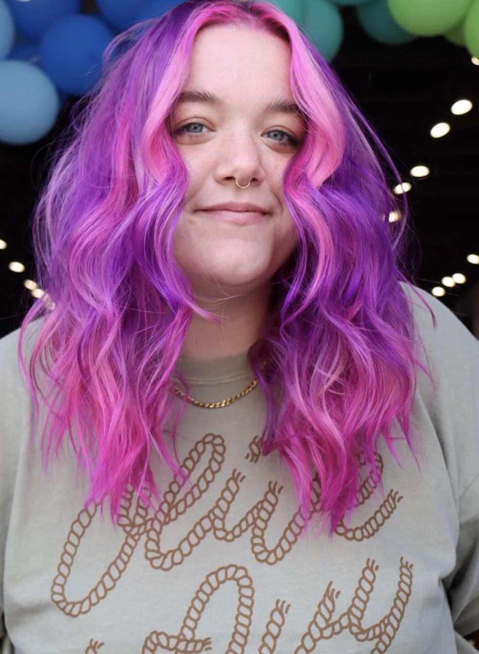 Young woman with vibrant pink and purple wavy hair, smiling, with colorful balloons in background.