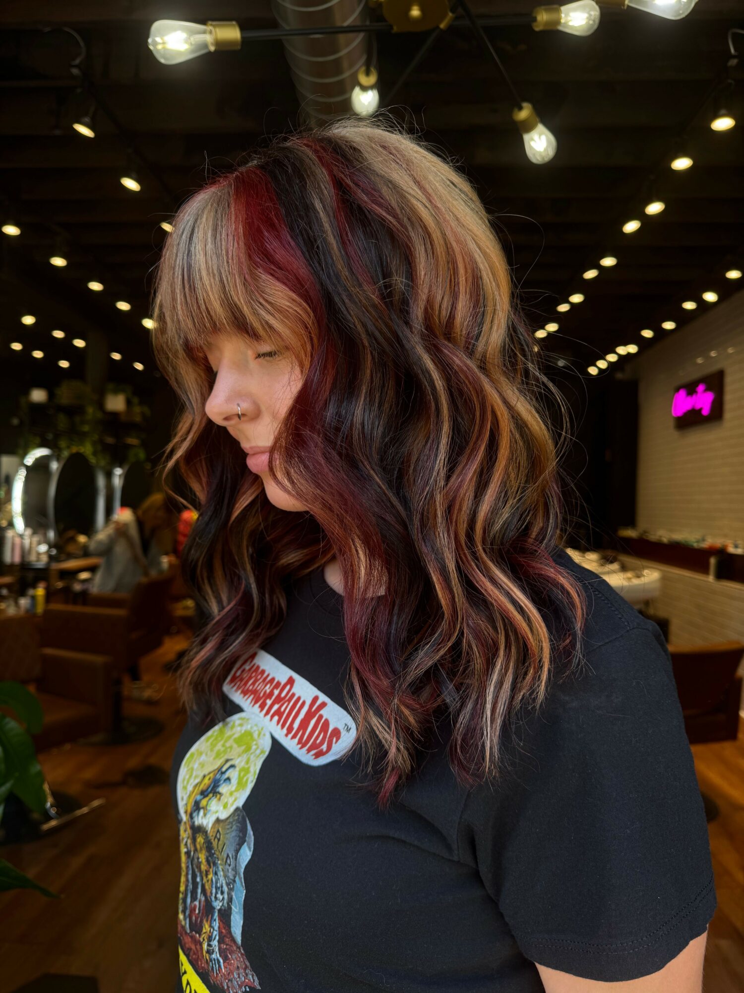 Side view of a woman with long, wavy hair, wearing a black shirt with a colorful graphic and text, in a dimly lit room.