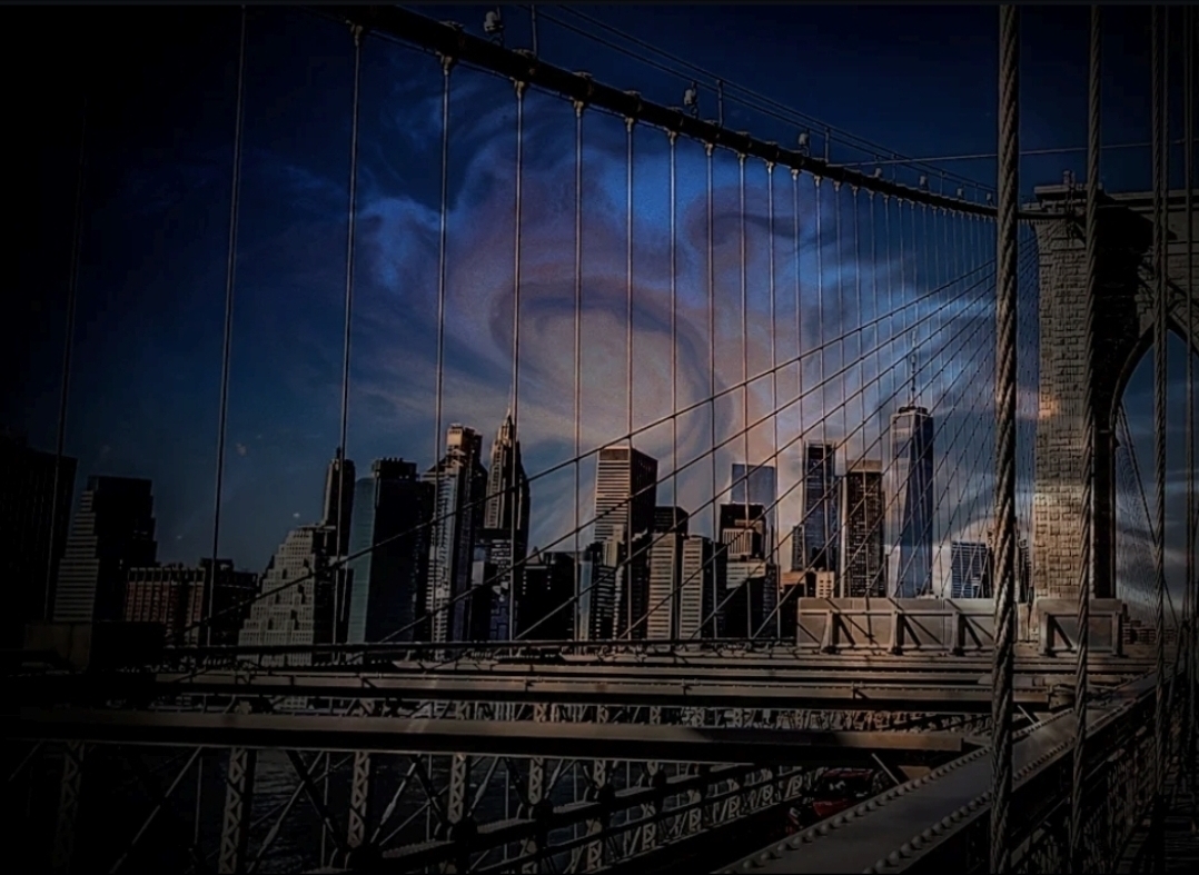 View of a city skyline with tall buildings seen through a bridge's suspension cables at dusk.