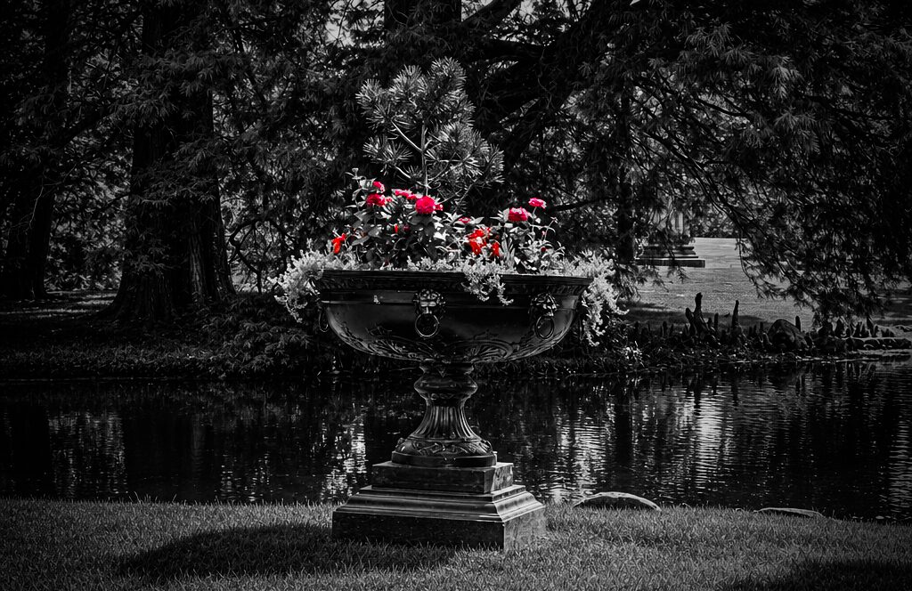 A large decorative stone urn filled with pink and red flowers, situated outdoors near a body of water, surrounded by trees.