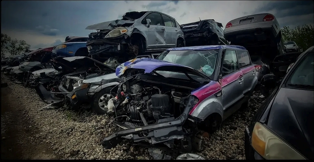 Multiple damaged cars stacked and scattered outdoors, with visible wreckage and debris, under cloudy sky.