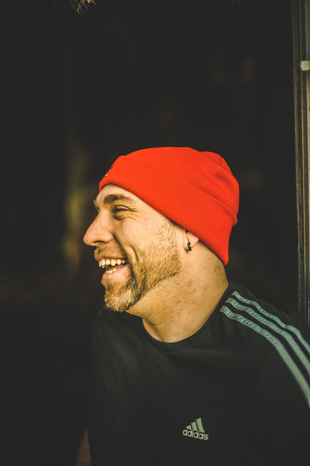 Side profile of a smiling man wearing a red beanie and black Adidas shirt, dark background