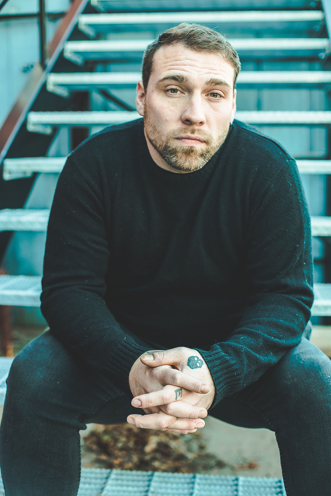 Man with short hair and beard sitting on stairs, wearing a black sweater, looking at camera.
