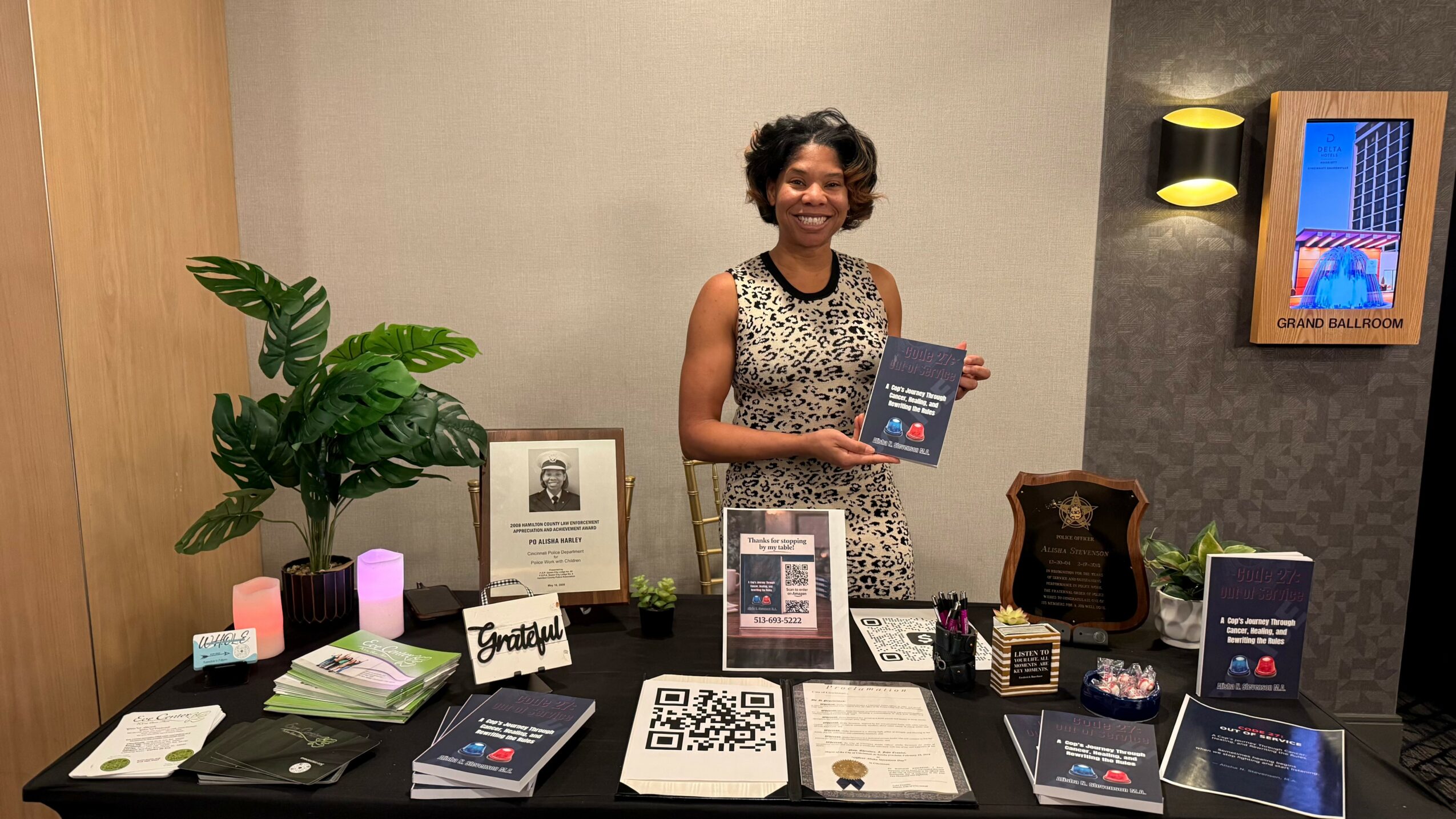 Woman standing behind a table with books and promotional materials, holding a book, in an indoor setting.
