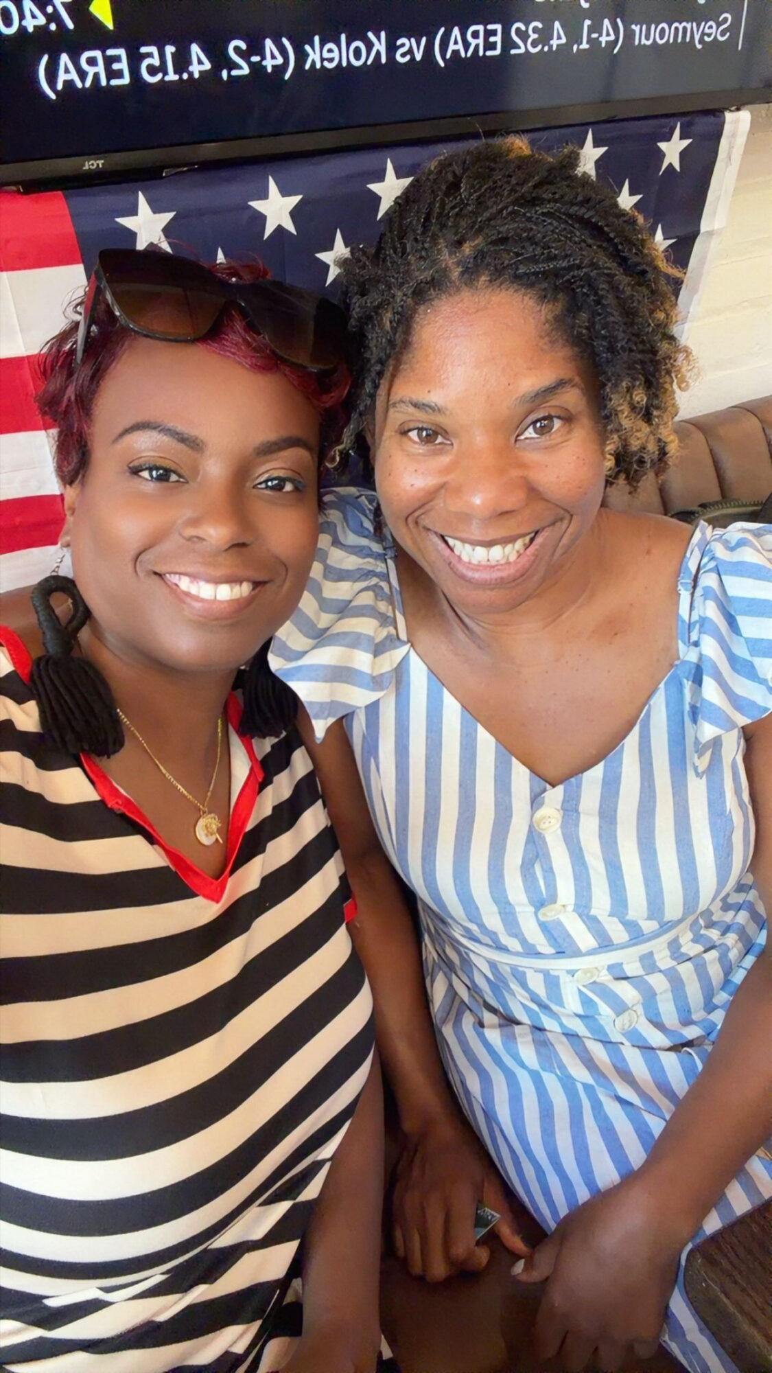 Two women smiling, one with sunglasses on her head, in front of an American flag background.