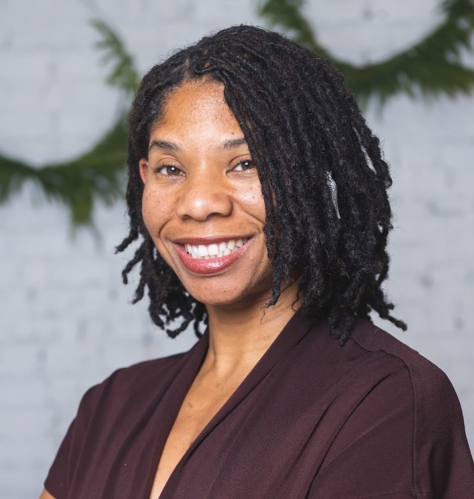 Smiling woman with shoulder-length dreadlocks wearing a dark top, standing in front of a white brick wall with greenery.