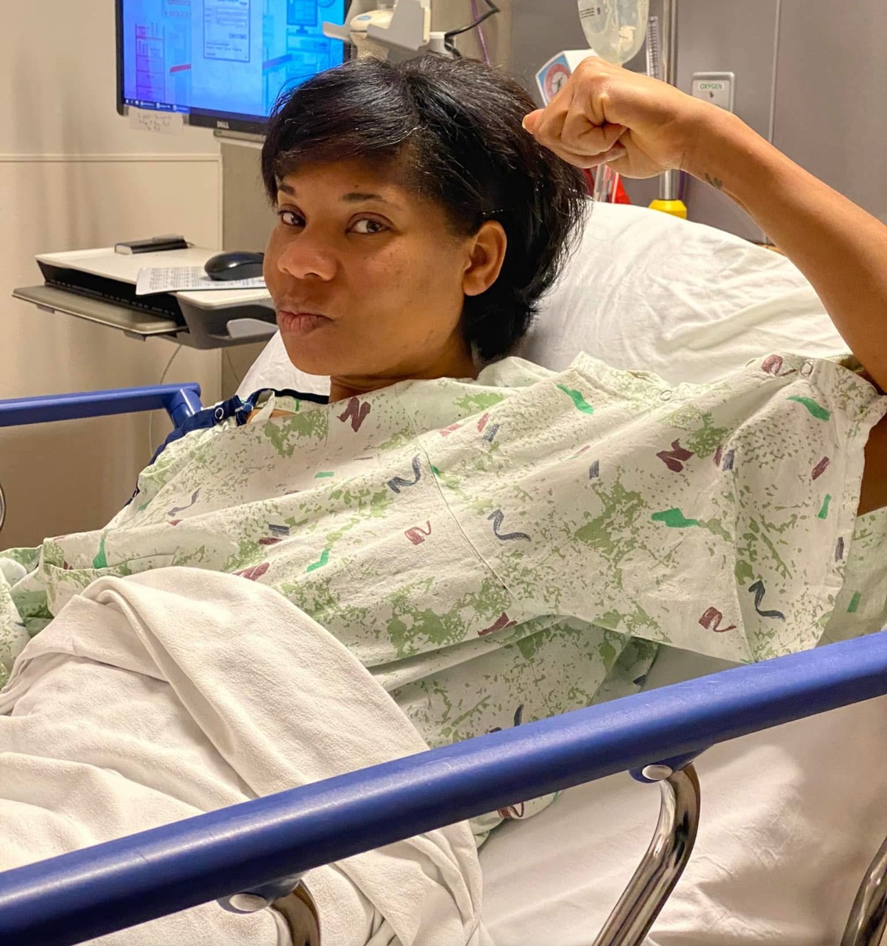 Woman in hospital bed flexing arm, wearing hospital gown, with medical equipment in background.