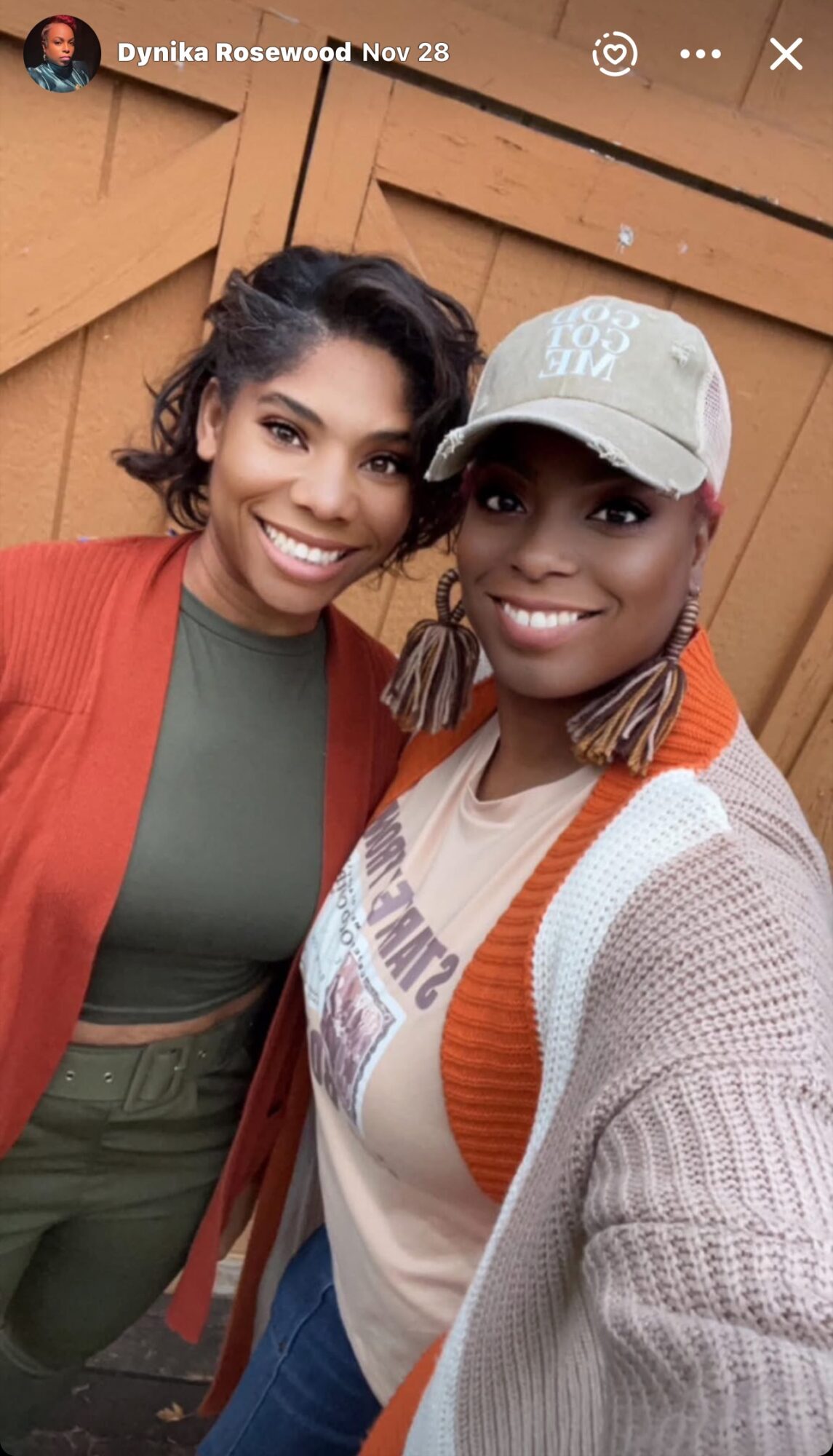 Two women smiling, standing close together in front of a wooden background, one wearing a red jacket and the other a white cap.
