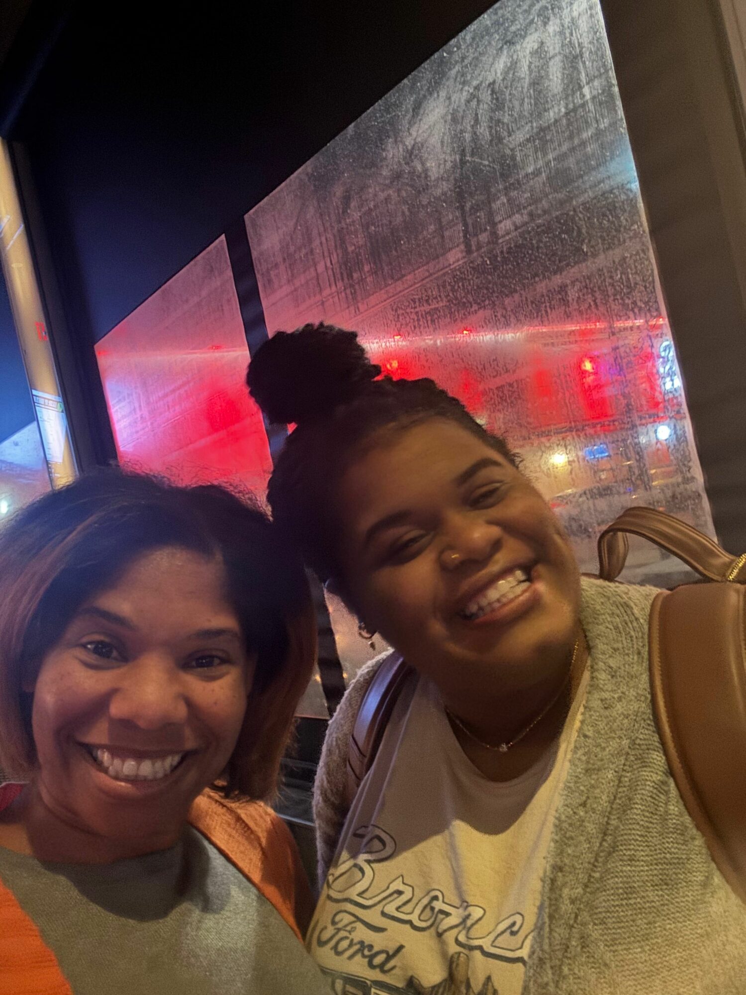 Two smiling women pose indoors with a reflective surface and colorful lights in the background.