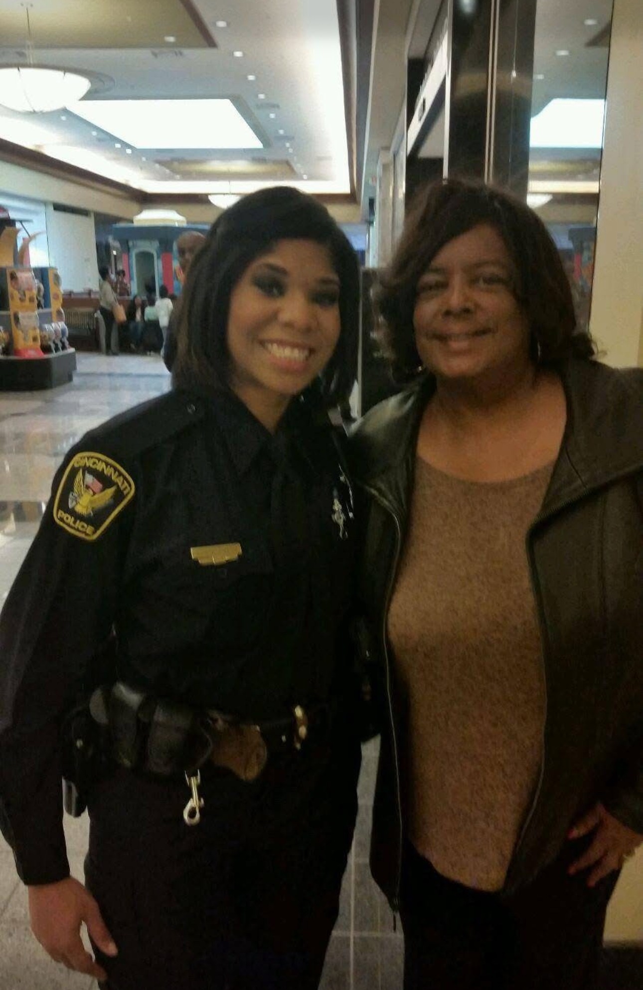 Two women standing together indoors, one in a police uniform and the other in casual clothing, smiling at the camera.