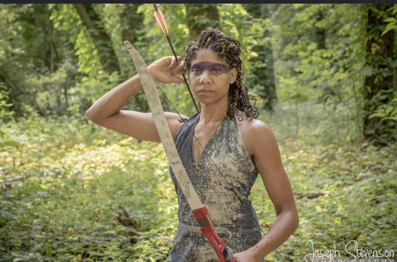 Woman in a forest holding a bow and arrow, wearing a sleeveless dress and glasses.