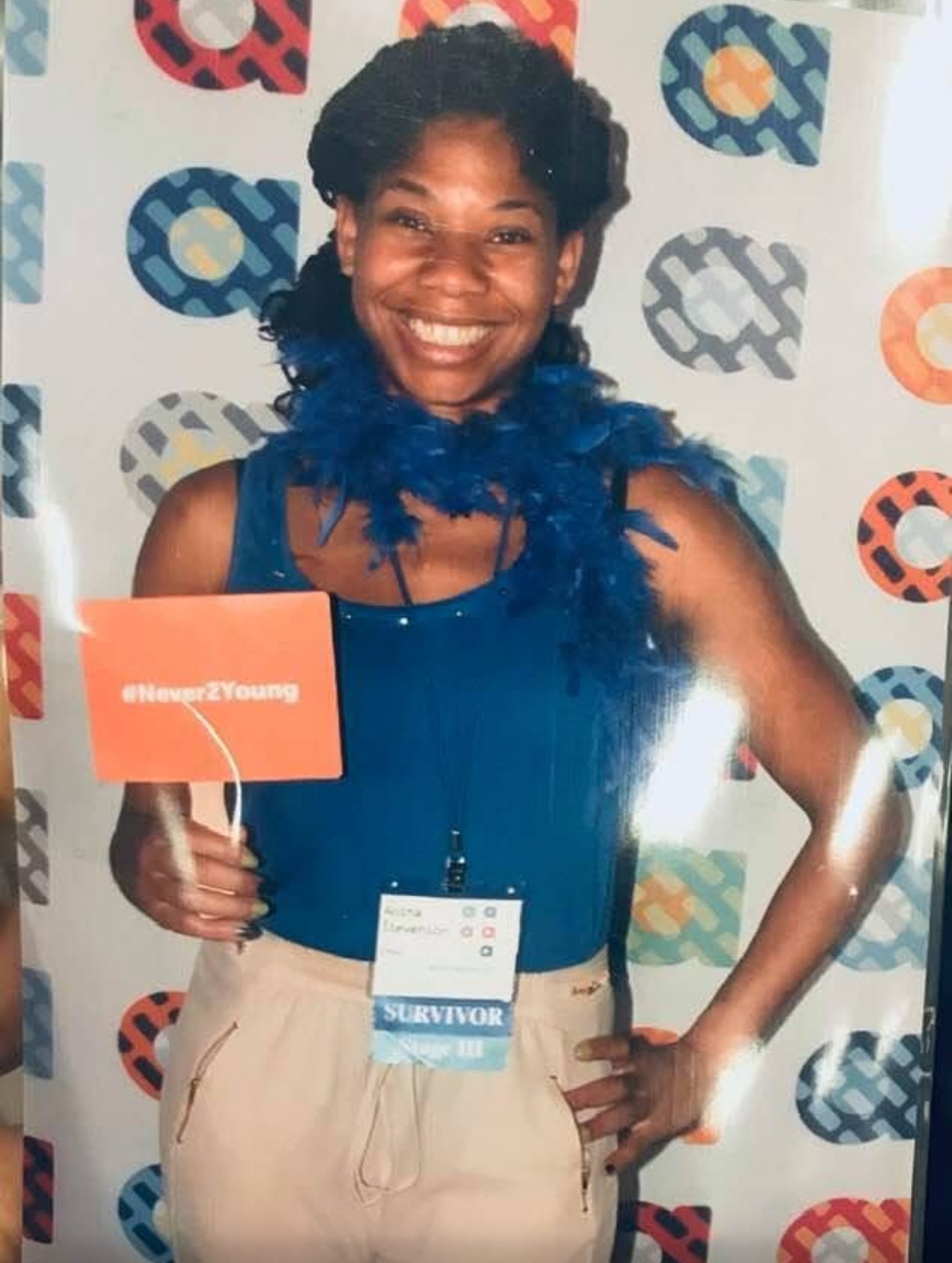 Woman smiling, holding a sign, wearing a blue feathered accessory, standing in front of a patterned backdrop.