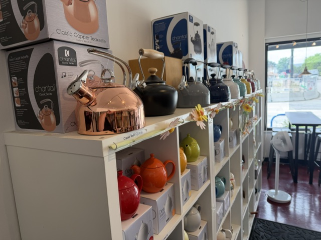 Shelves with colorful teapots and kitchen appliances inside a store, with a window and seating area outside.