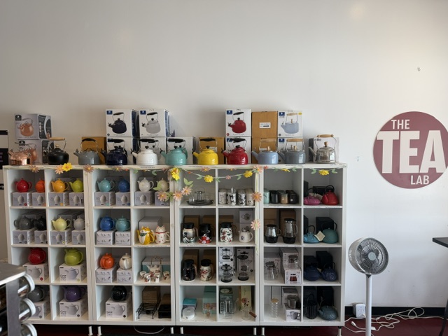 Shelves filled with colorful tea accessories and boxes, with a fan on the floor and a sign reading 'The Tea Lab' on the wall.