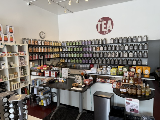Interior of a tea shop with shelves of tea containers, a table with tea products, and a wall sign reading 'The Tea'.