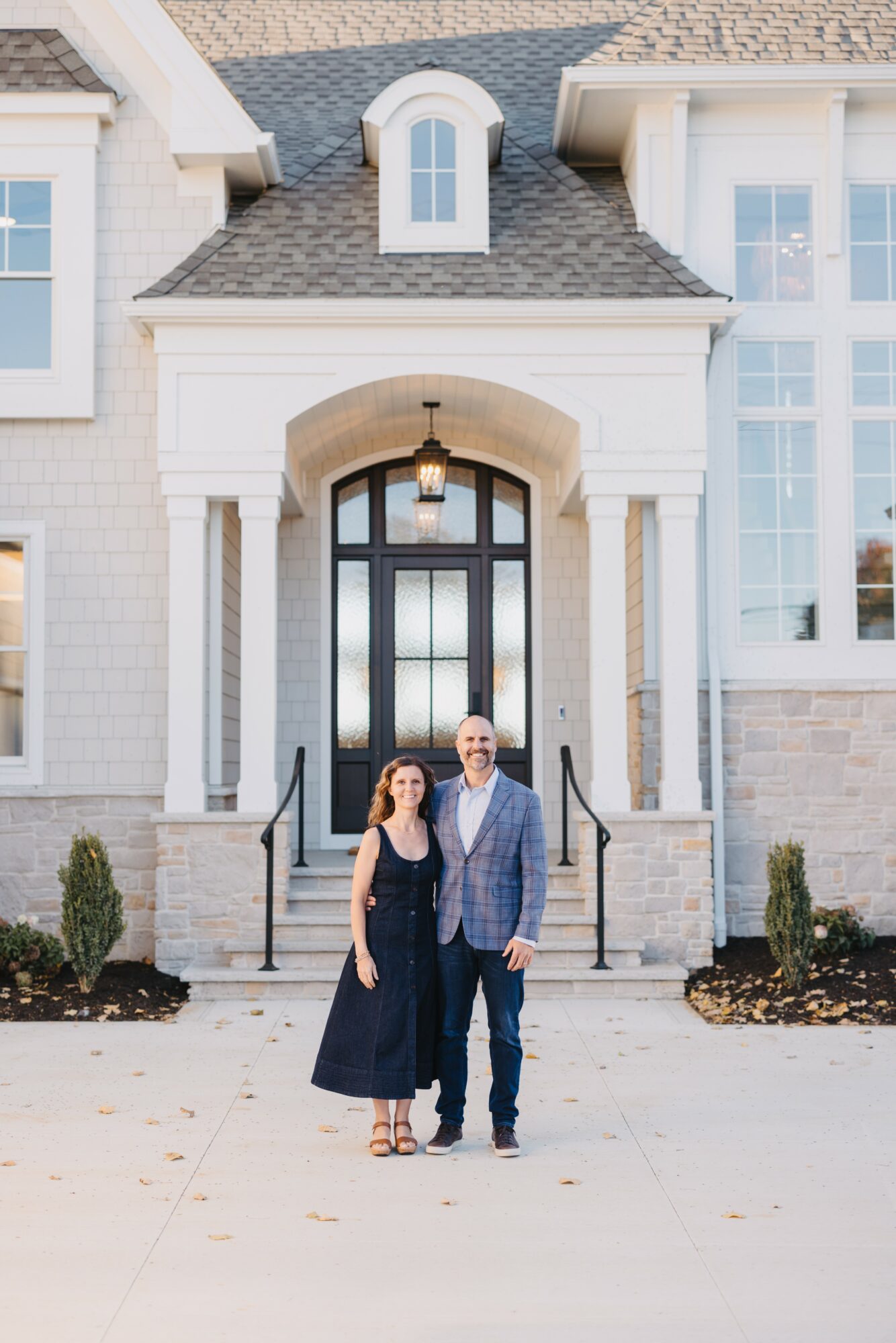 A man and woman stand on steps in front of a large house with white columns and a black door.