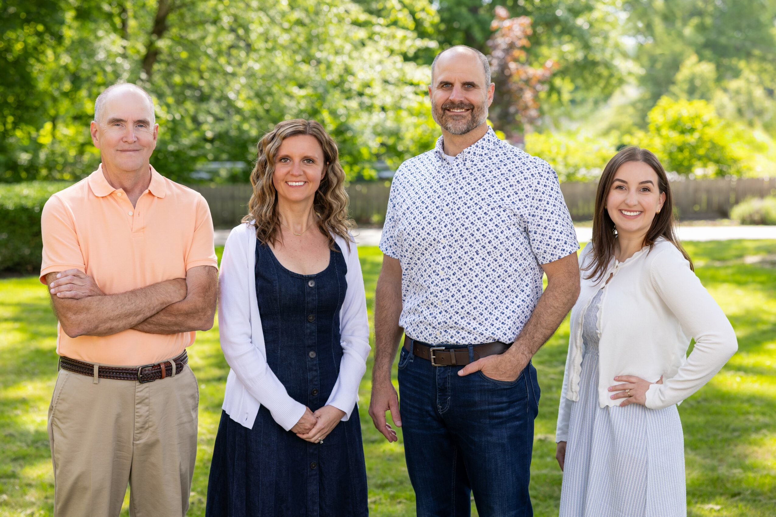 Four people standing outdoors in a park with trees and grass, smiling at the camera.