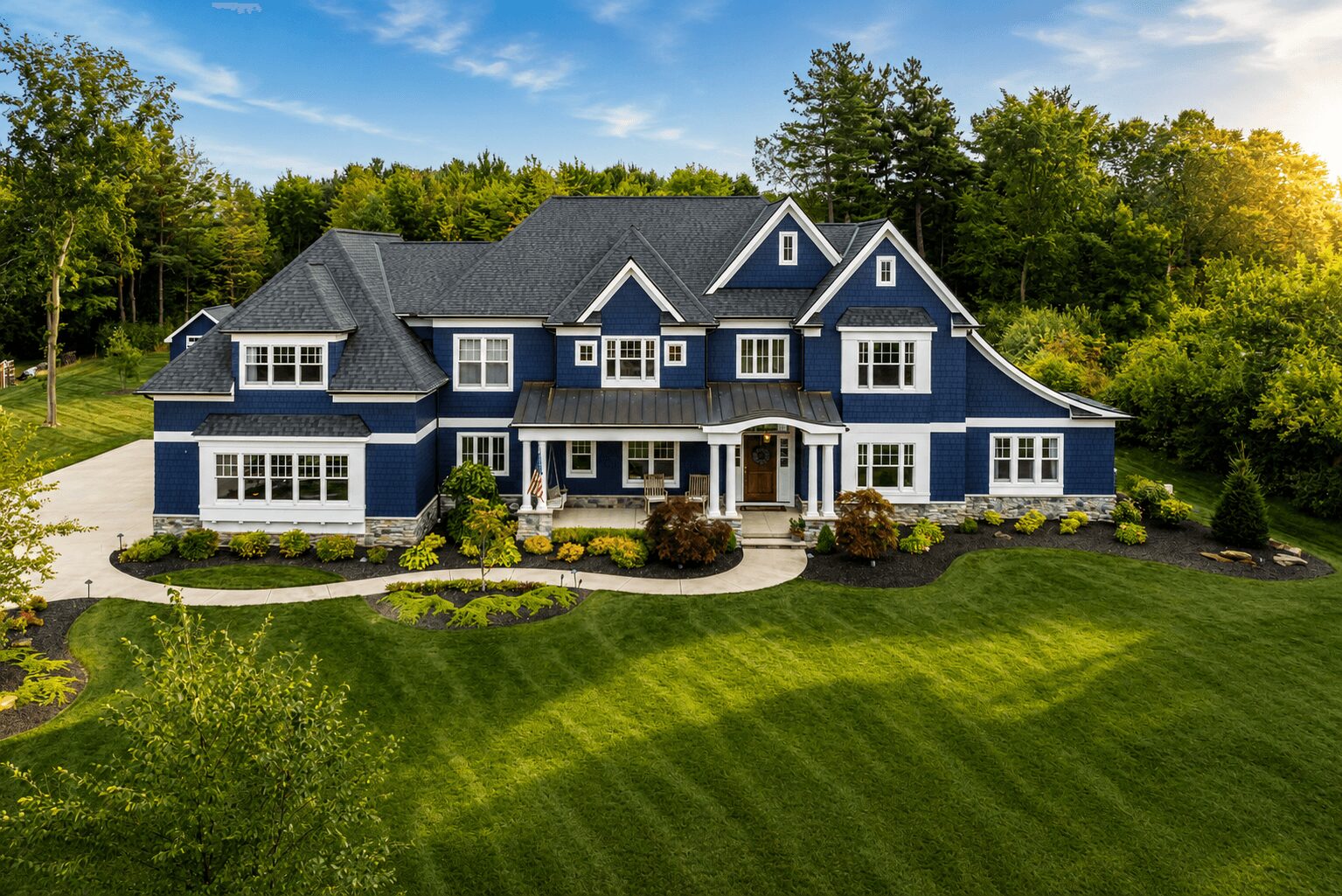 Large blue house with multiple gables, surrounded by green lawn and trees, under a clear sky with sunlight.