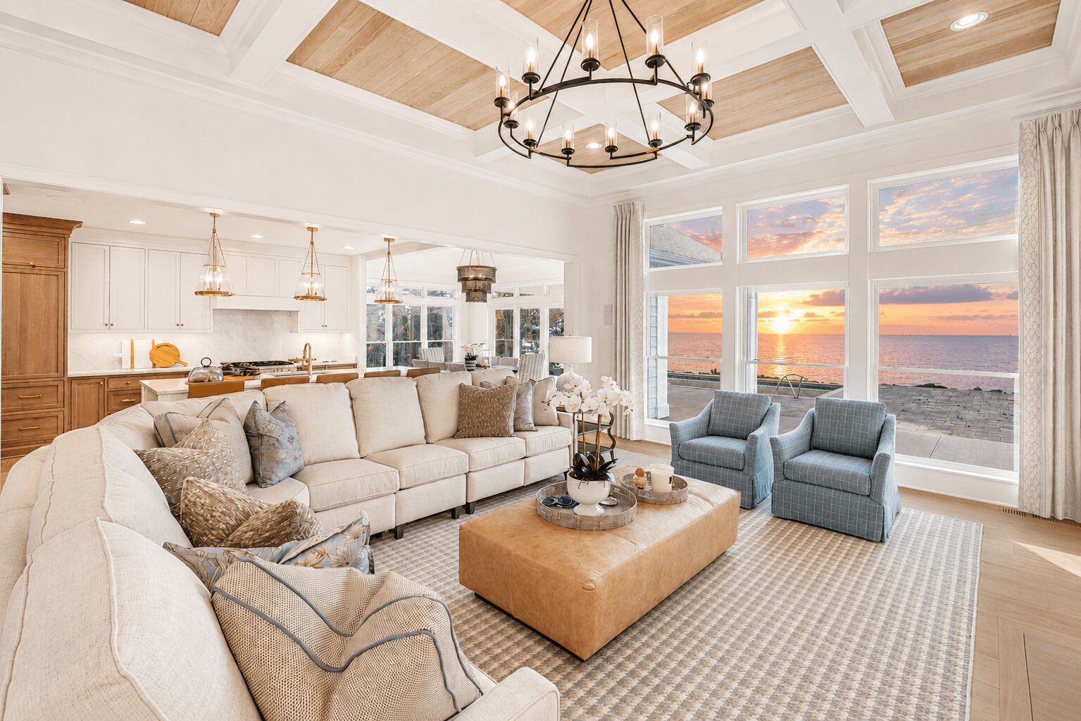 Living room with large beige sectional sofa, two blue armchairs, a beige ottoman, and a view of sunset over water through large windows.