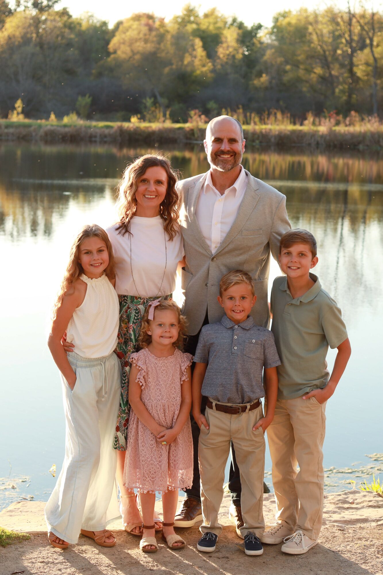 Family of seven standing by a lake with trees in the background, smiling, dressed casually, sunny day.