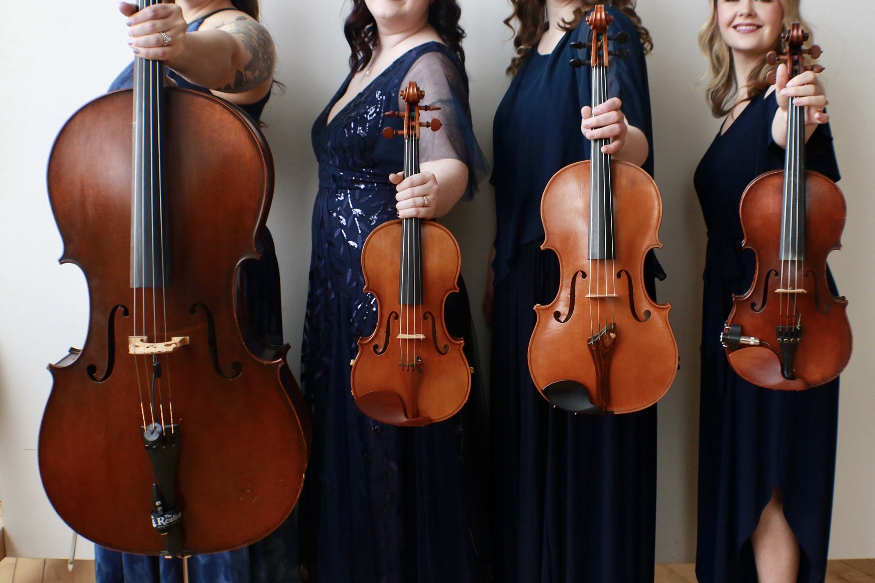 Four women holding string instruments, including a double bass and violins, dressed in dark blue dresses, standing side by side.