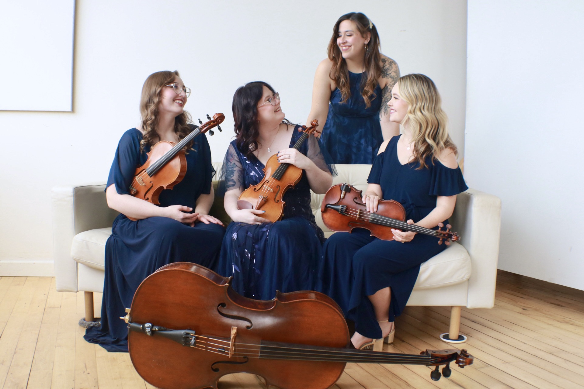 Five women in matching dark blue dresses with violins and a cello, sitting and standing on a white sofa in a room.