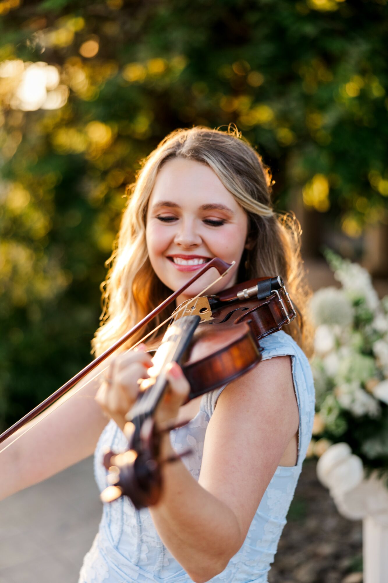 Young woman playing violin outdoors, smiling, with trees and flowers in background.