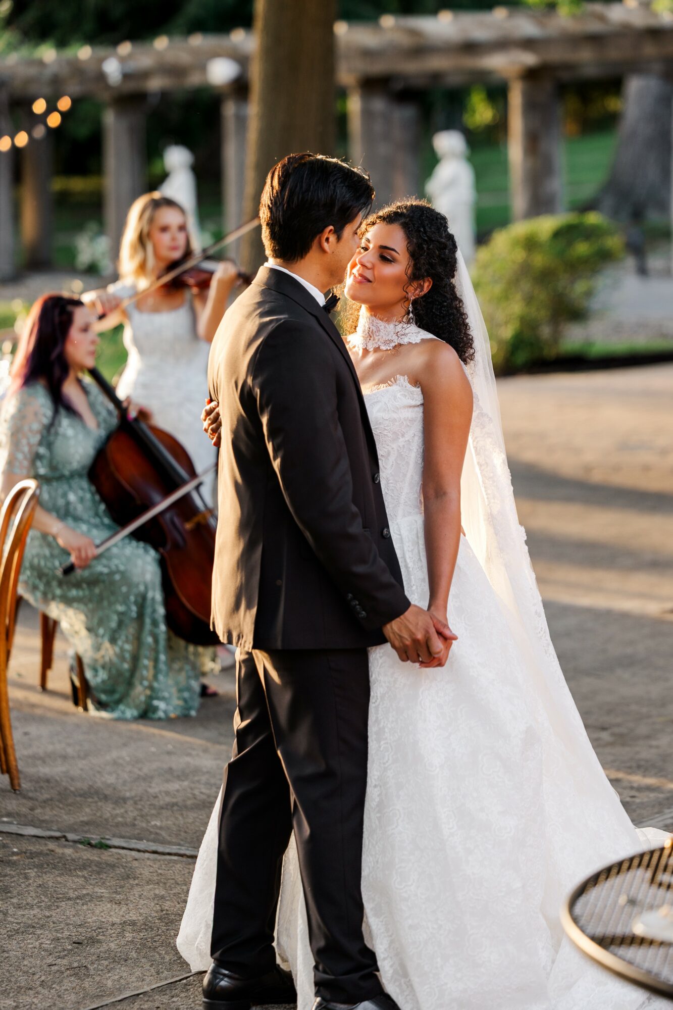 Bride and groom holding hands and facing each other outdoors, with musicians playing in the background.