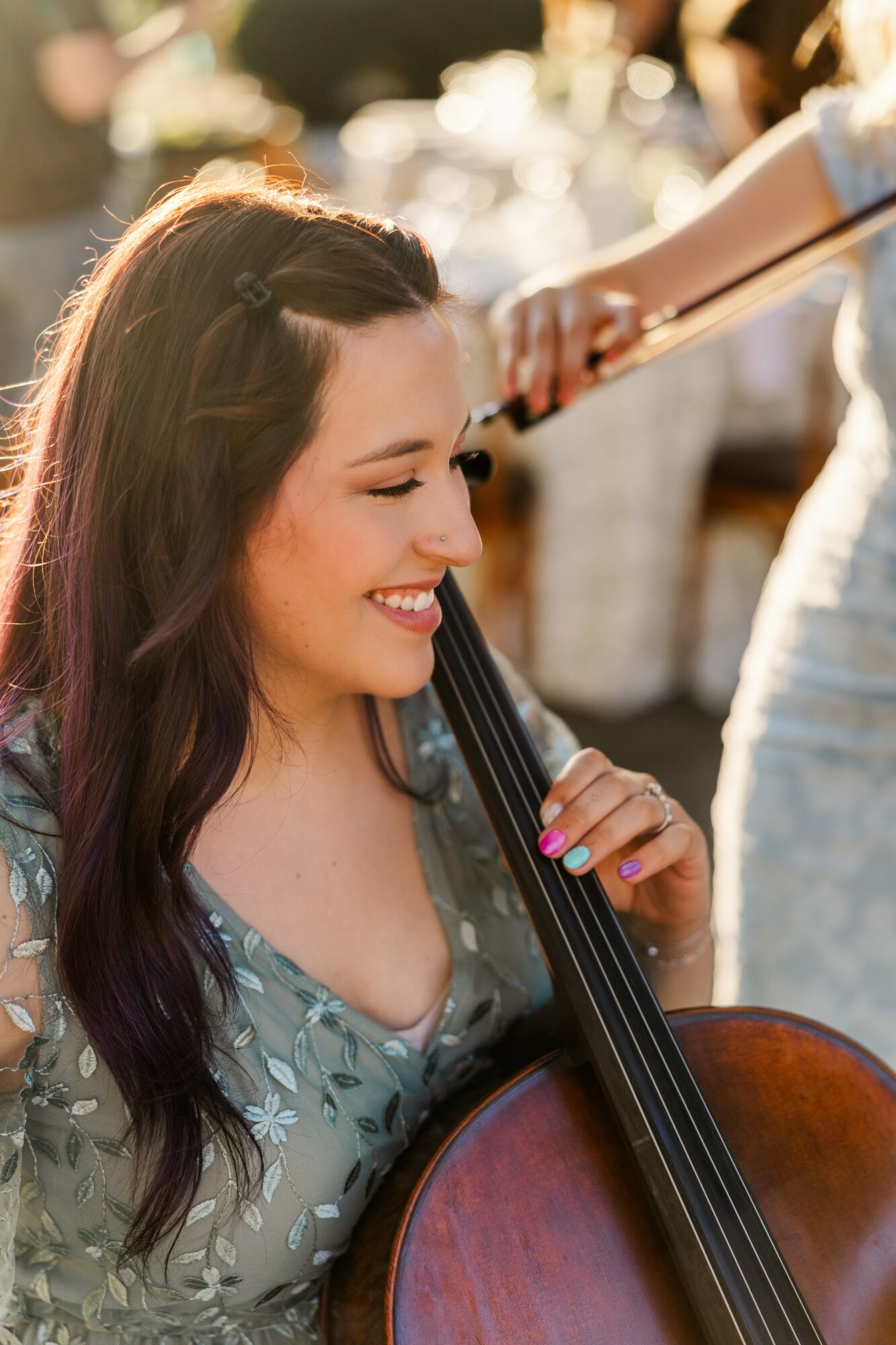 Woman with long dark hair smiling, holding a cello, in a bright outdoor setting.