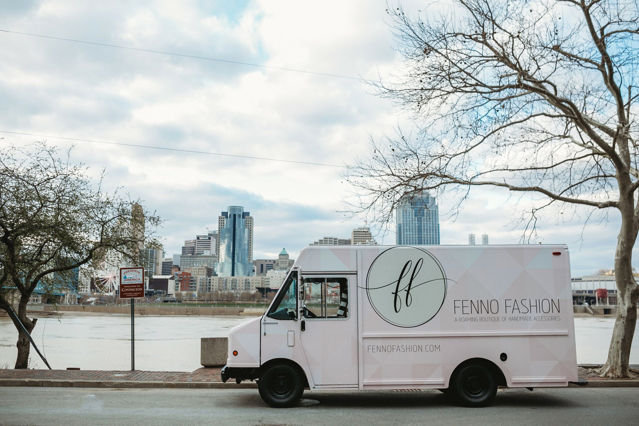 White delivery truck with Fenno Fashion logo parked near river, city skyline in background, leafless trees, cloudy sky.