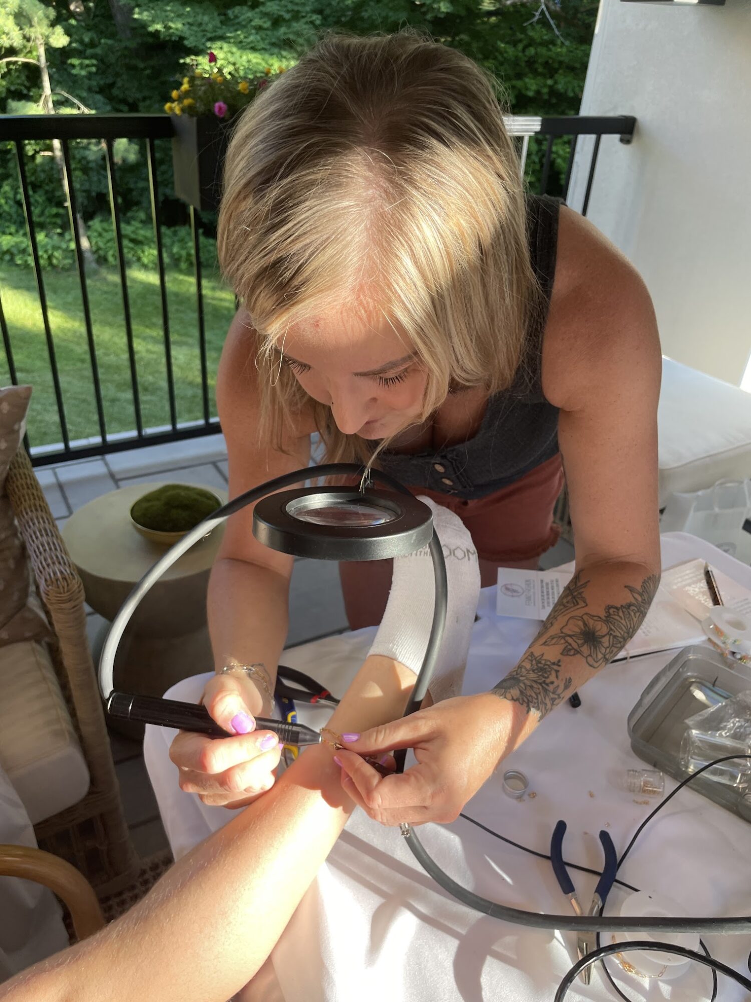 Person with blonde hair examining a hand under a magnifying device outdoors, with a tattooed arm and a table nearby.
