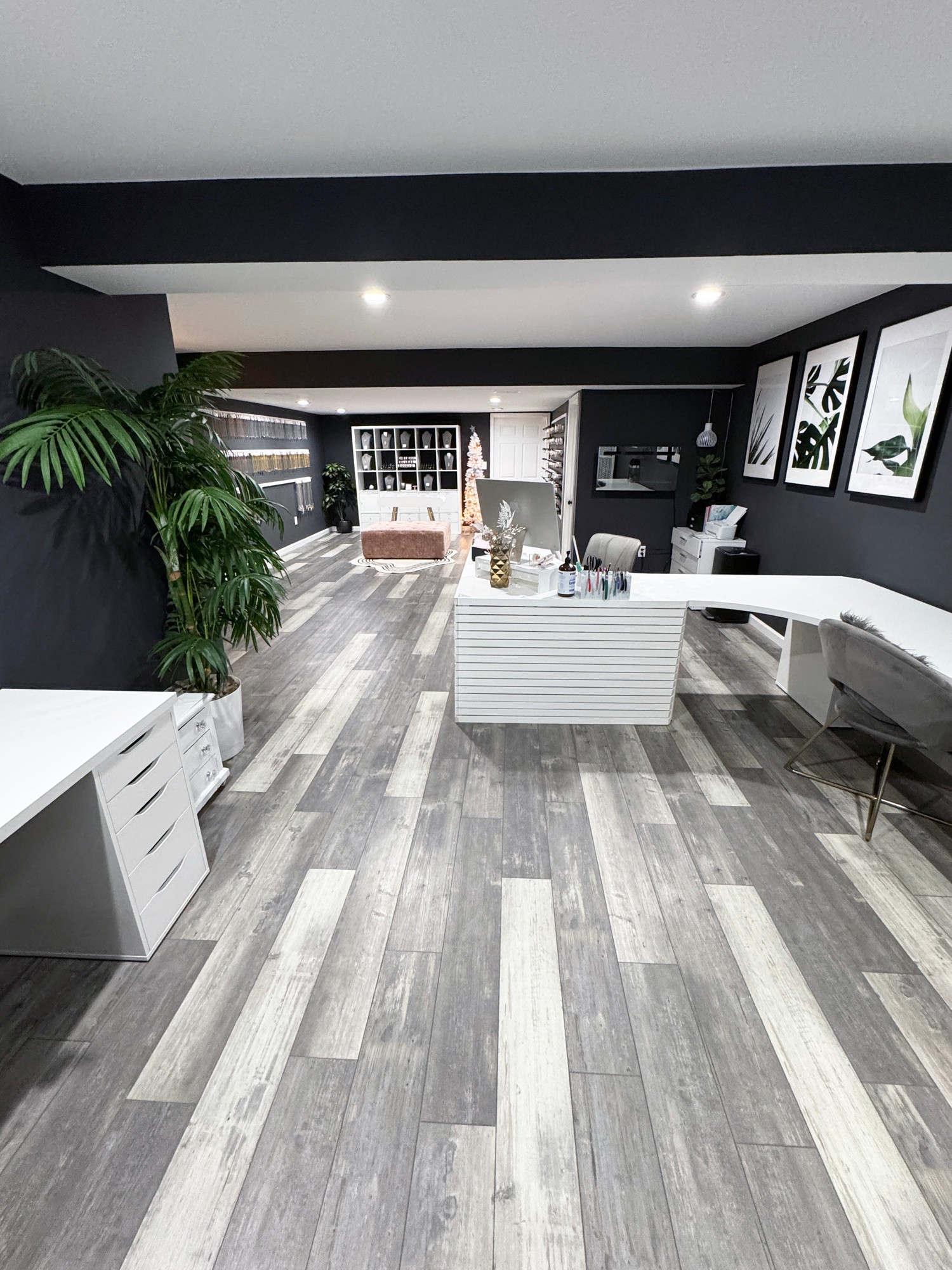 Modern office space with white desks, black walls, large windows, and framed artwork, featuring a gray wood floor and a large plant.